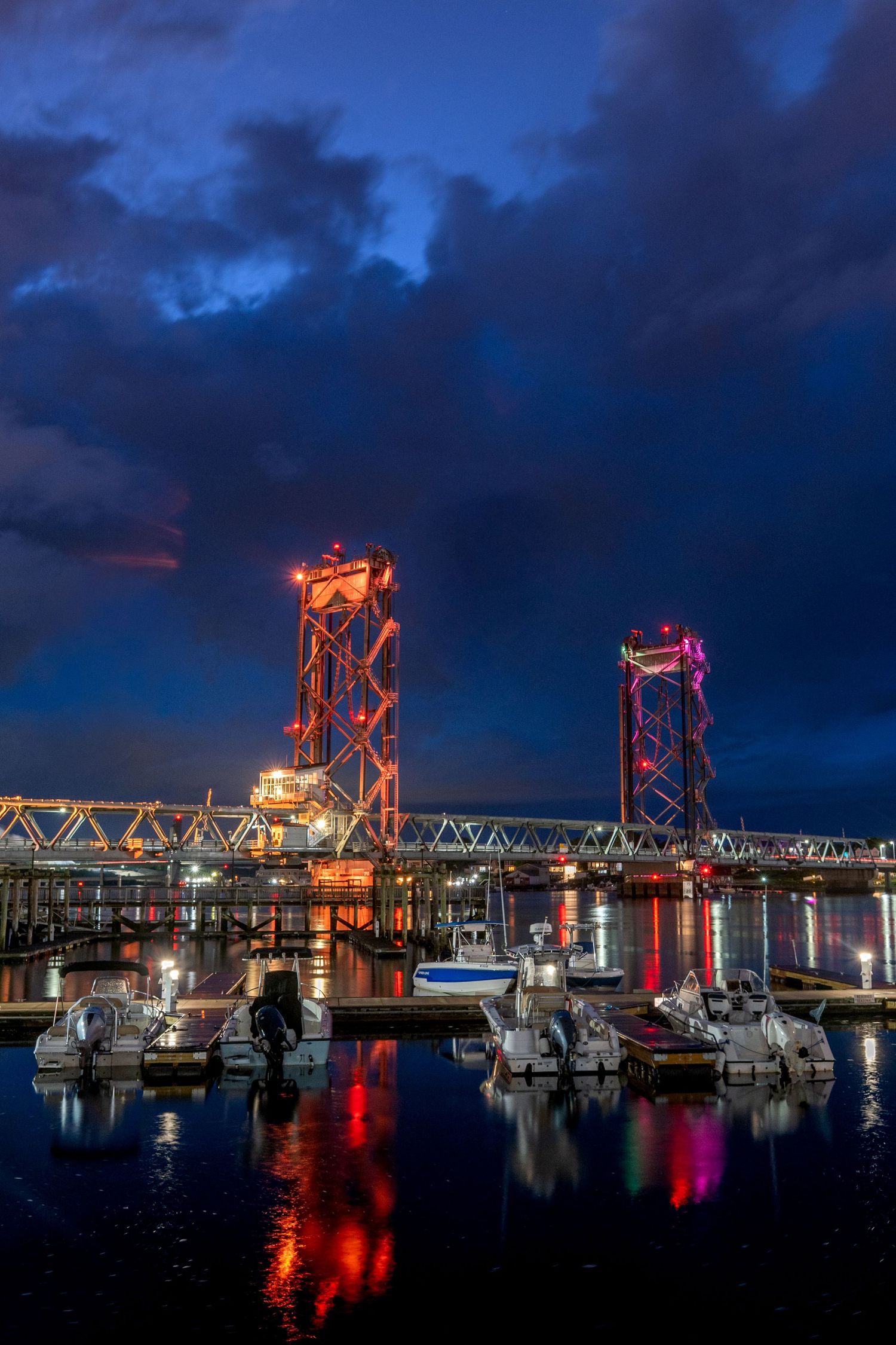 Illuminated lift bridge with colorful reflections on calm harbor waters at dusk, boats docked in foreground under dramatic cloudy sky.