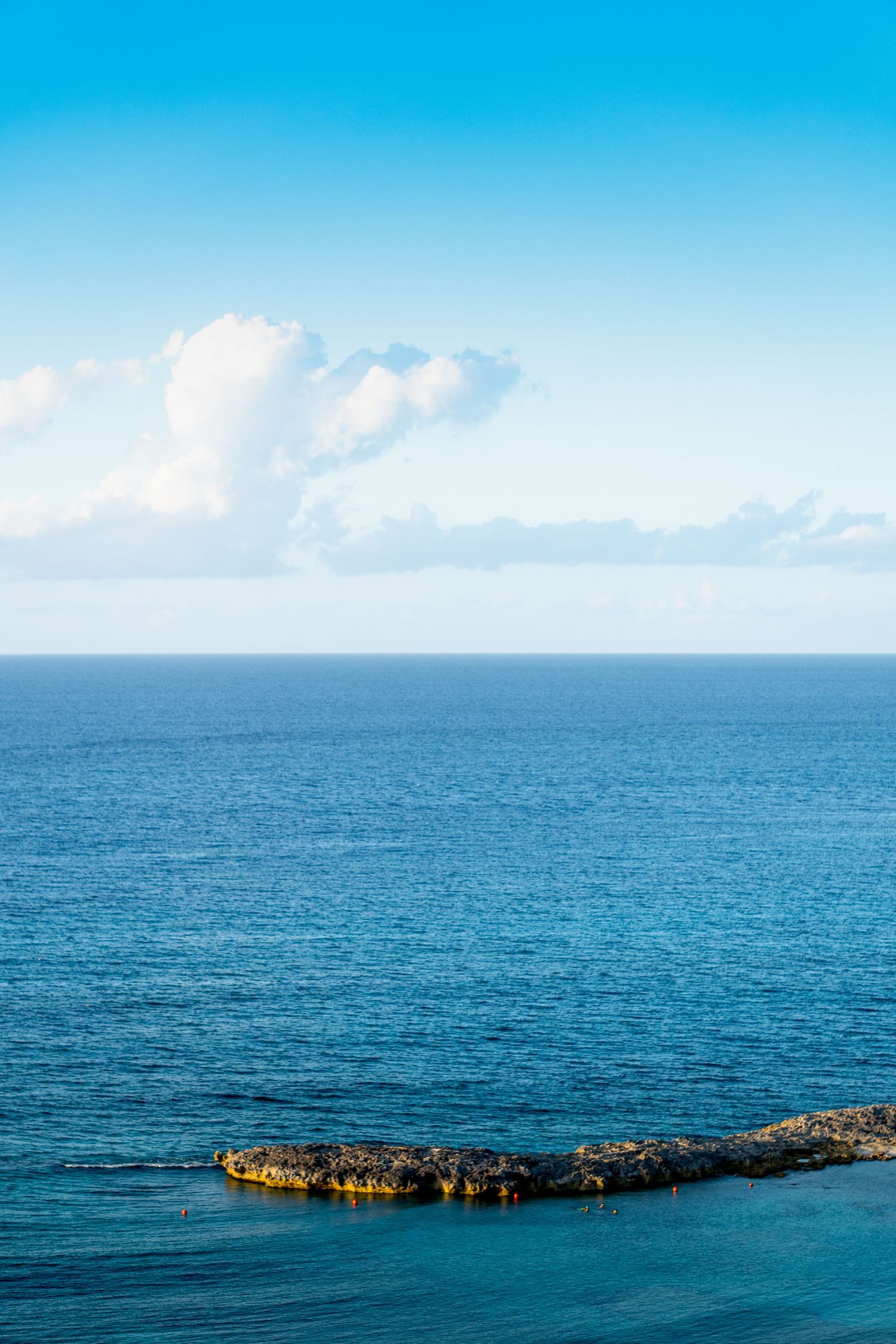 Panoramic view of a calm blue ocean with a rocky outcrop visible against the horizon under bright blue skies.