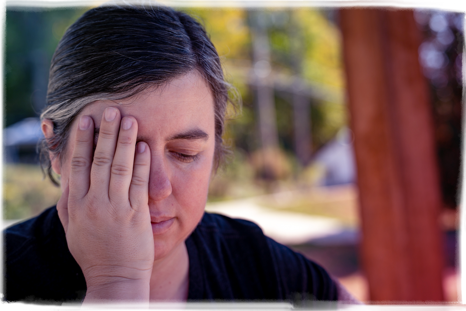 Person covering their face with one hand in an outdoor setting with blurred background.