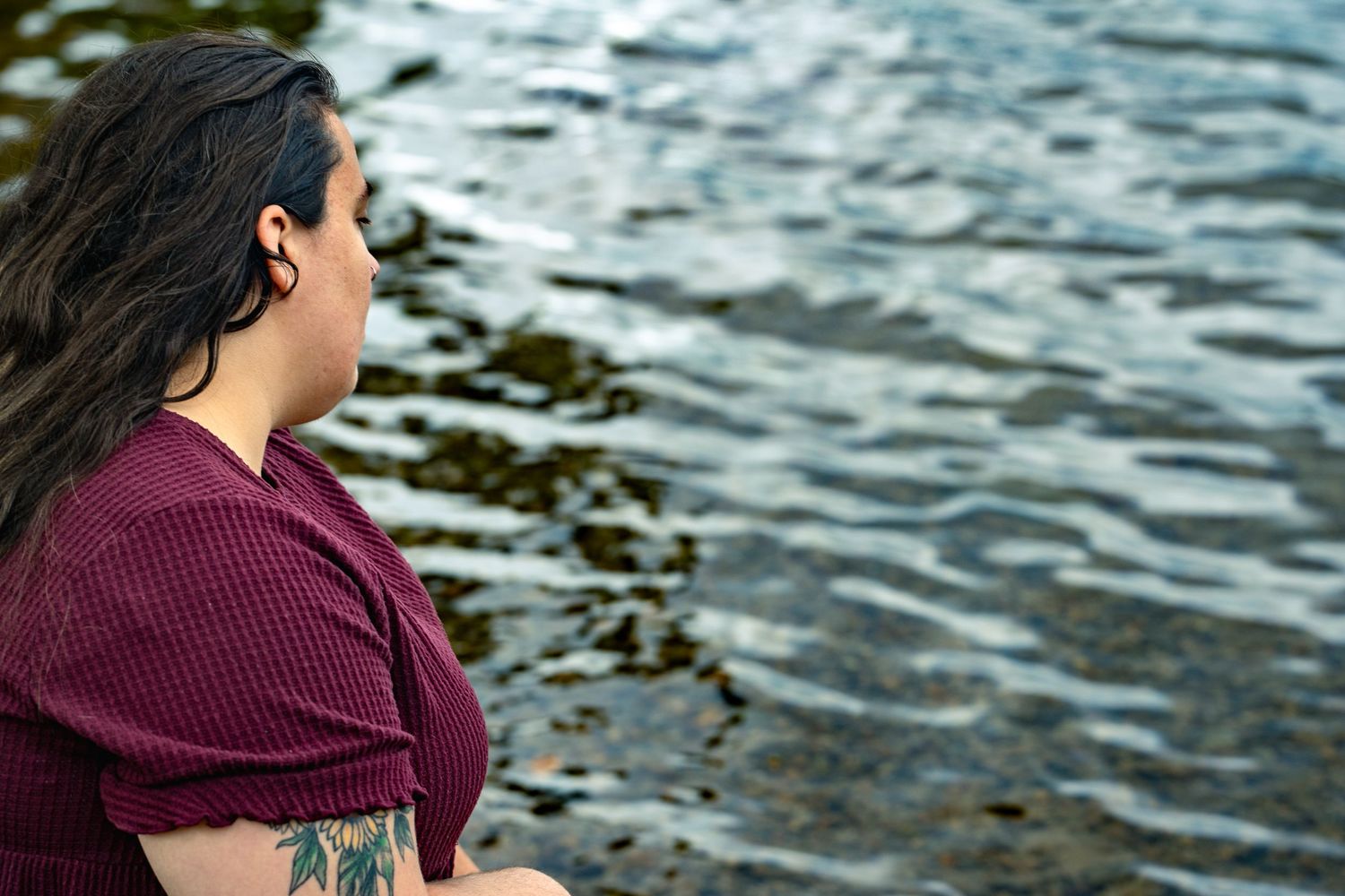 Figure in maroon top sits contemplatively by rippling water under moody skies.
