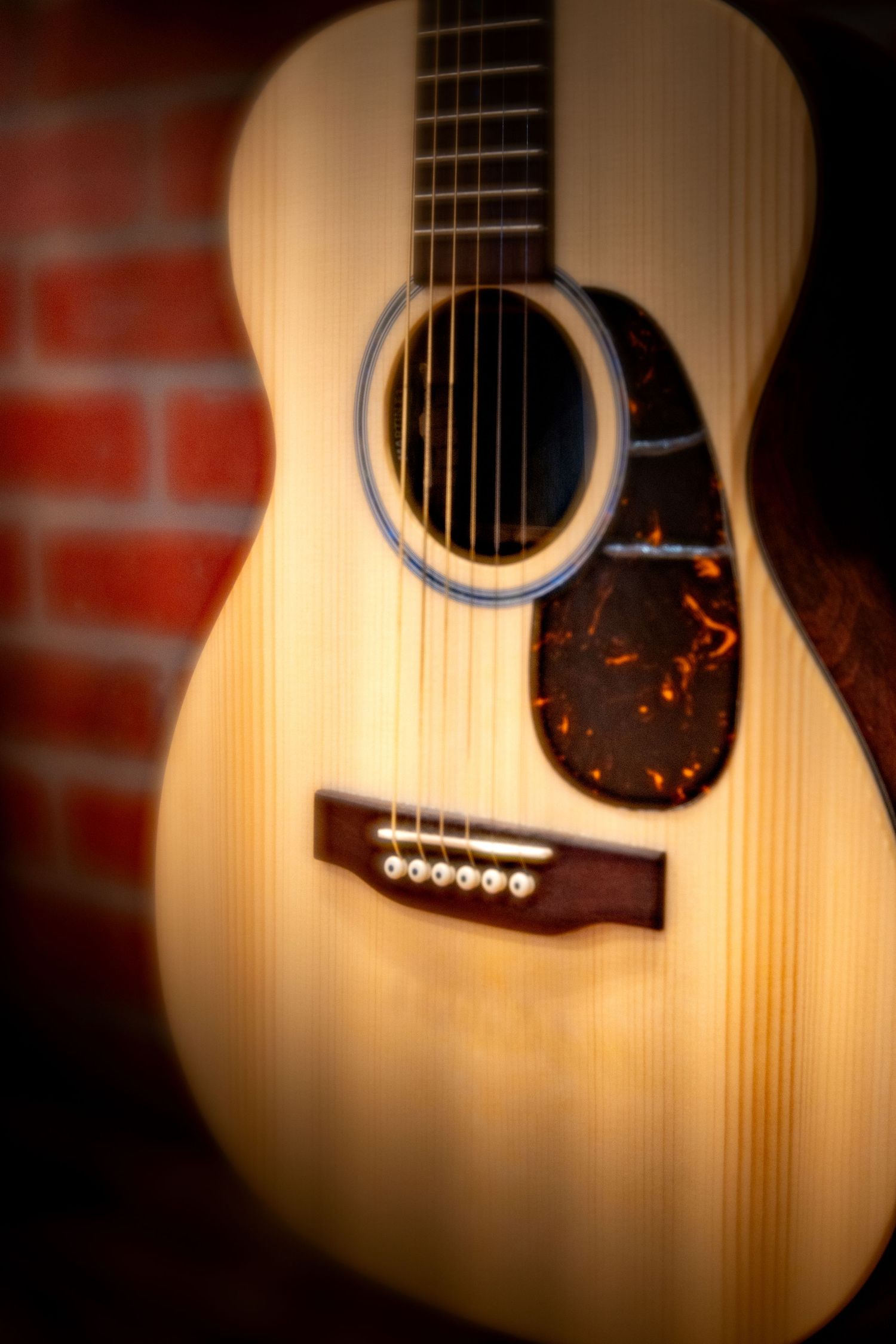 Close-up of a natural wood acoustic guitar with tortoiseshell pickguard leaning against a red brick wall.