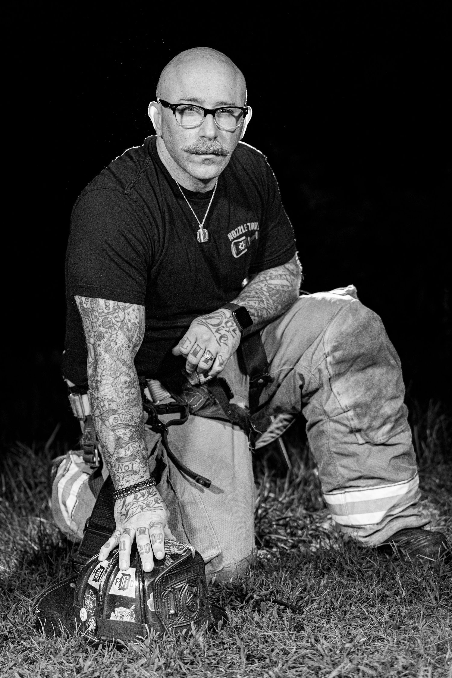 Black and white photo of someone with tattoos and glasses crouching down in grass at night.