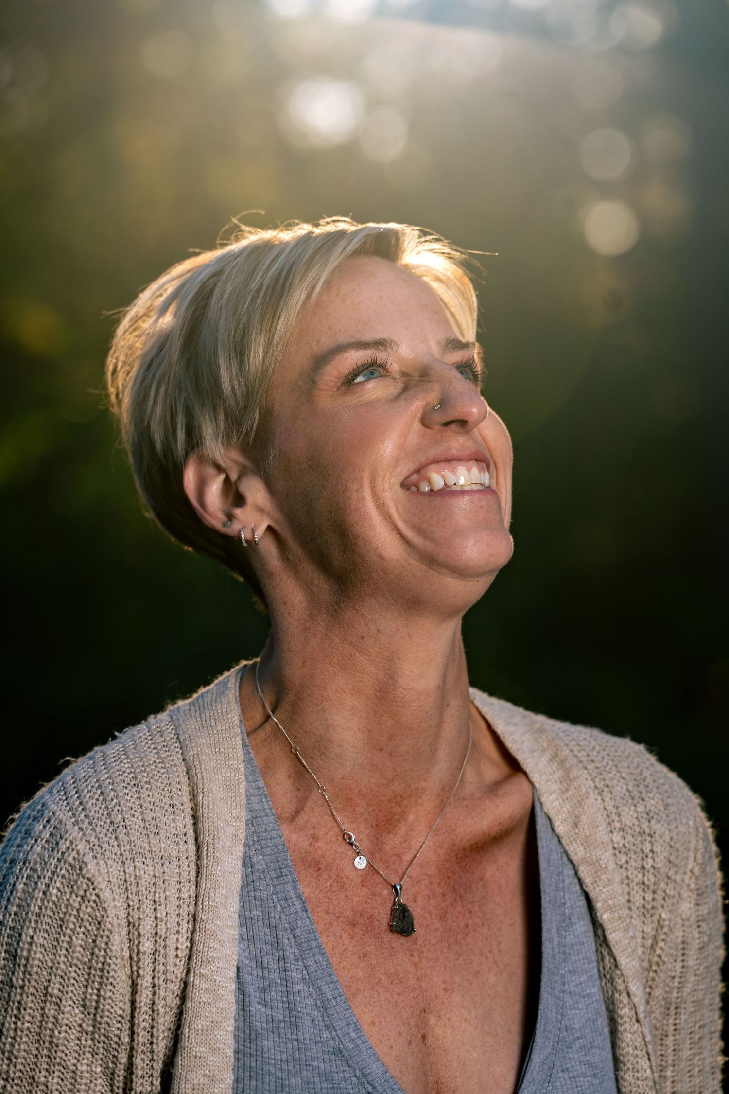 A person wearing a gray cardigan and blue top smiles warmly while looking up at natural sunlit bokeh in the background.