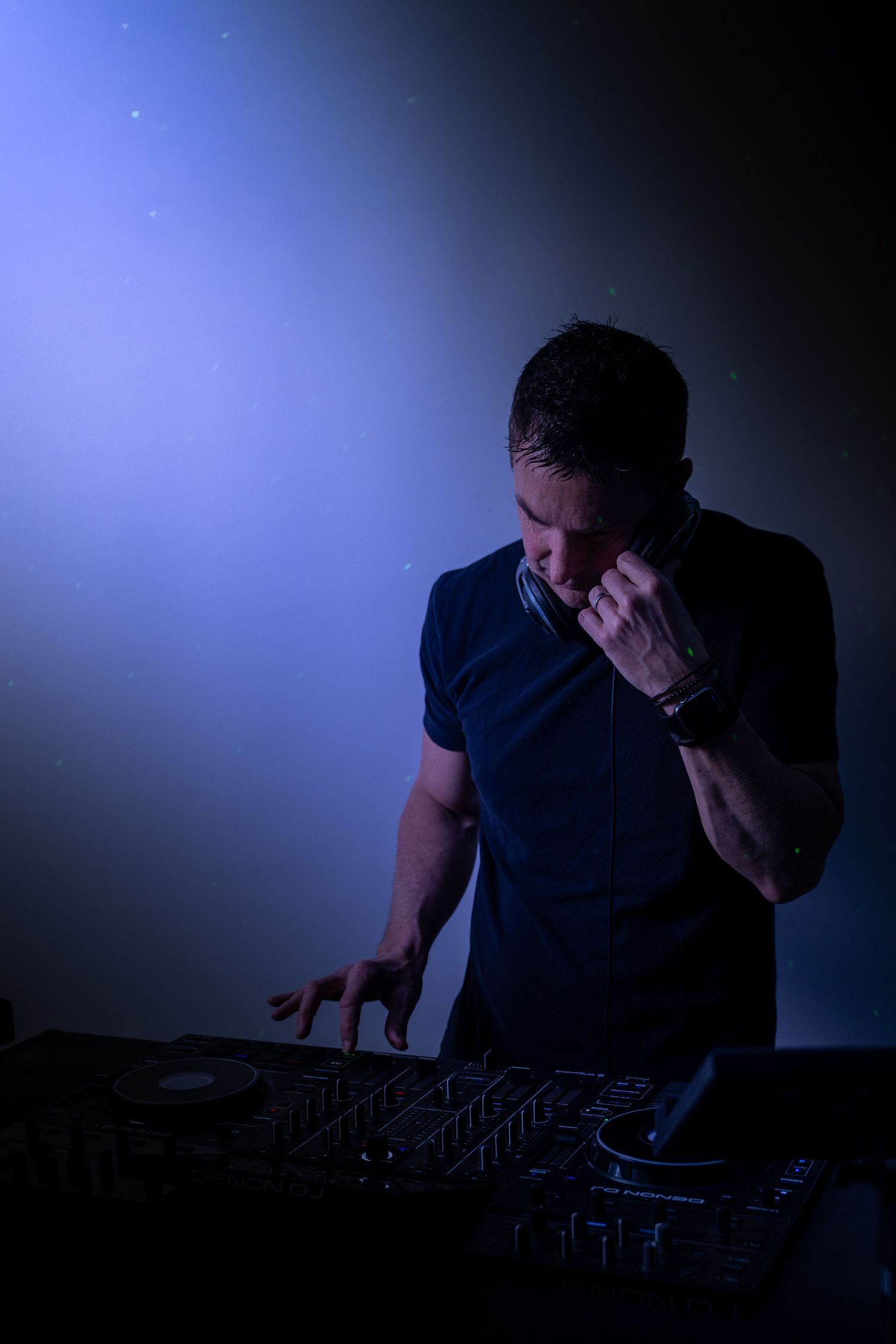 A DJ performing at a nightclub, holding headphones to his ear while mixing tracks on a professional DJ controller under blue stage lighting.