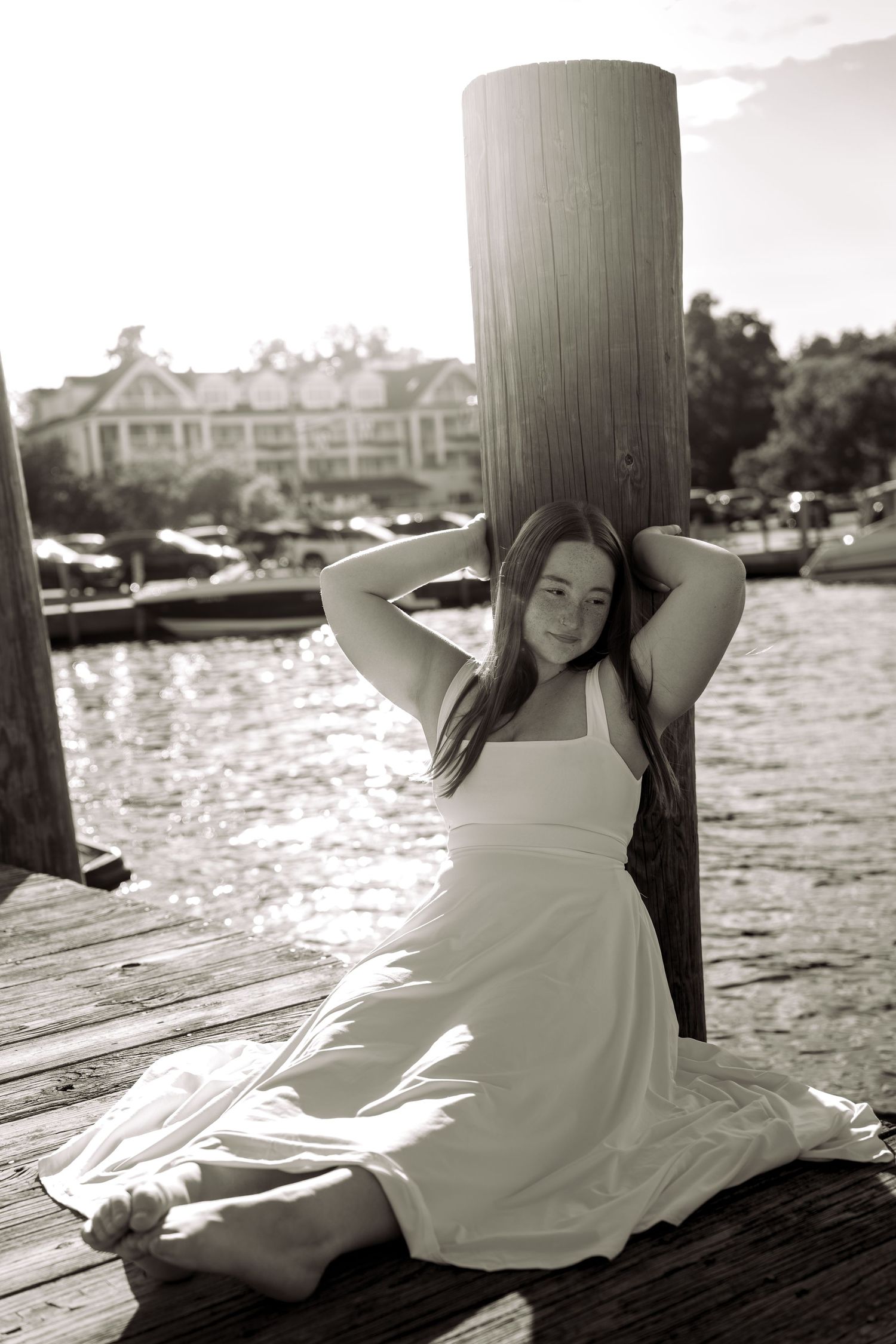Black and white photo of a young woman in a white dress sitting on a wooden dock by the water.