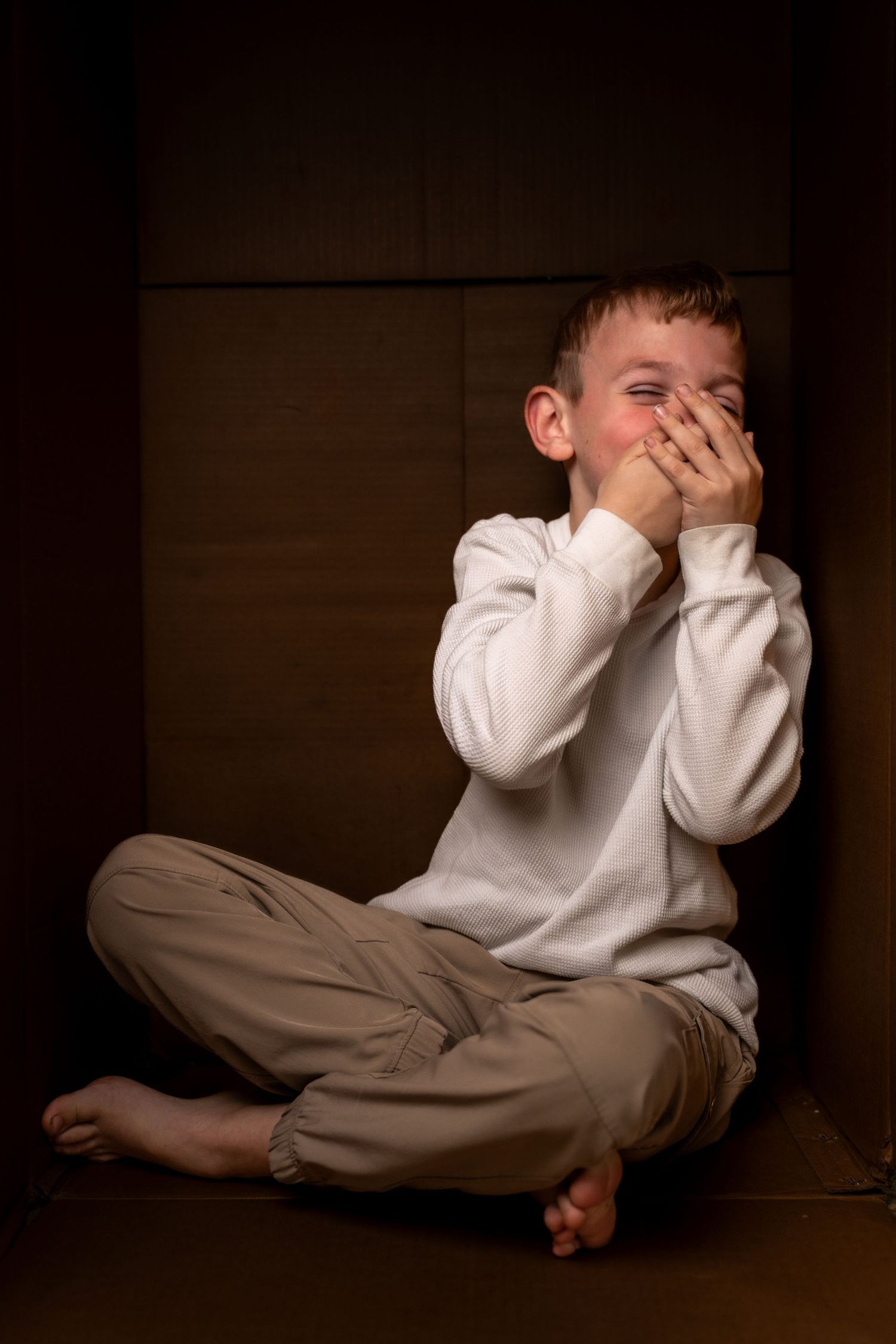 Young barefoot boy sitting cross-legged in dark cardboard box, covering face with hands, appearing distressed.