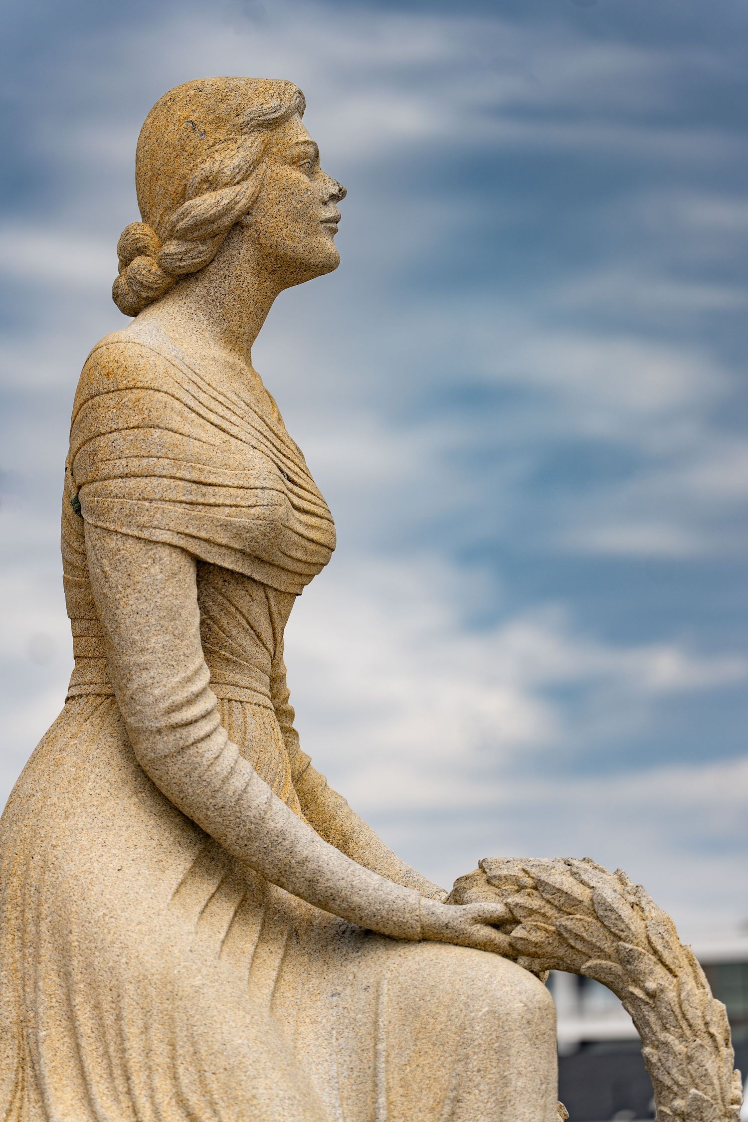 Stone statue of an elegant woman in a flowing dress, gazing upward against a cloudy blue sky.