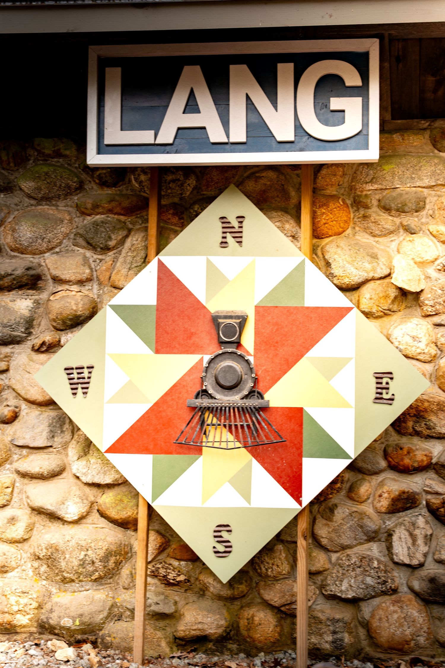 Colorful geometric quilt barn sign with vintage train motif below Lang station sign on stone wall.