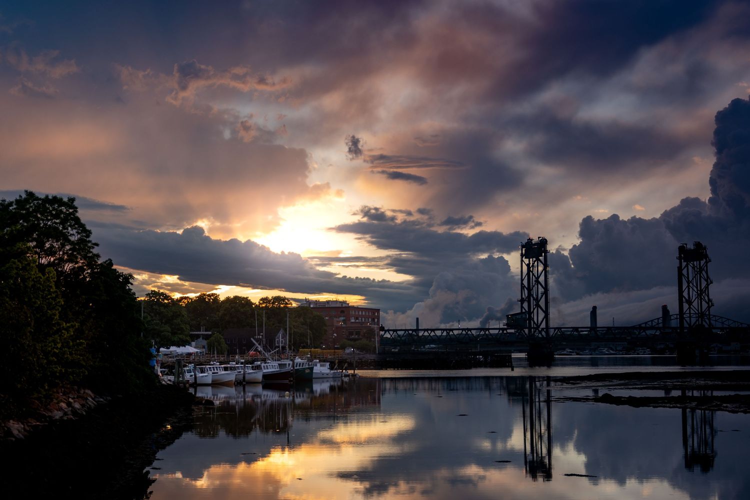 Dramatic sunset over a marina with a lift bridge, stormy clouds reflecting on calm water.