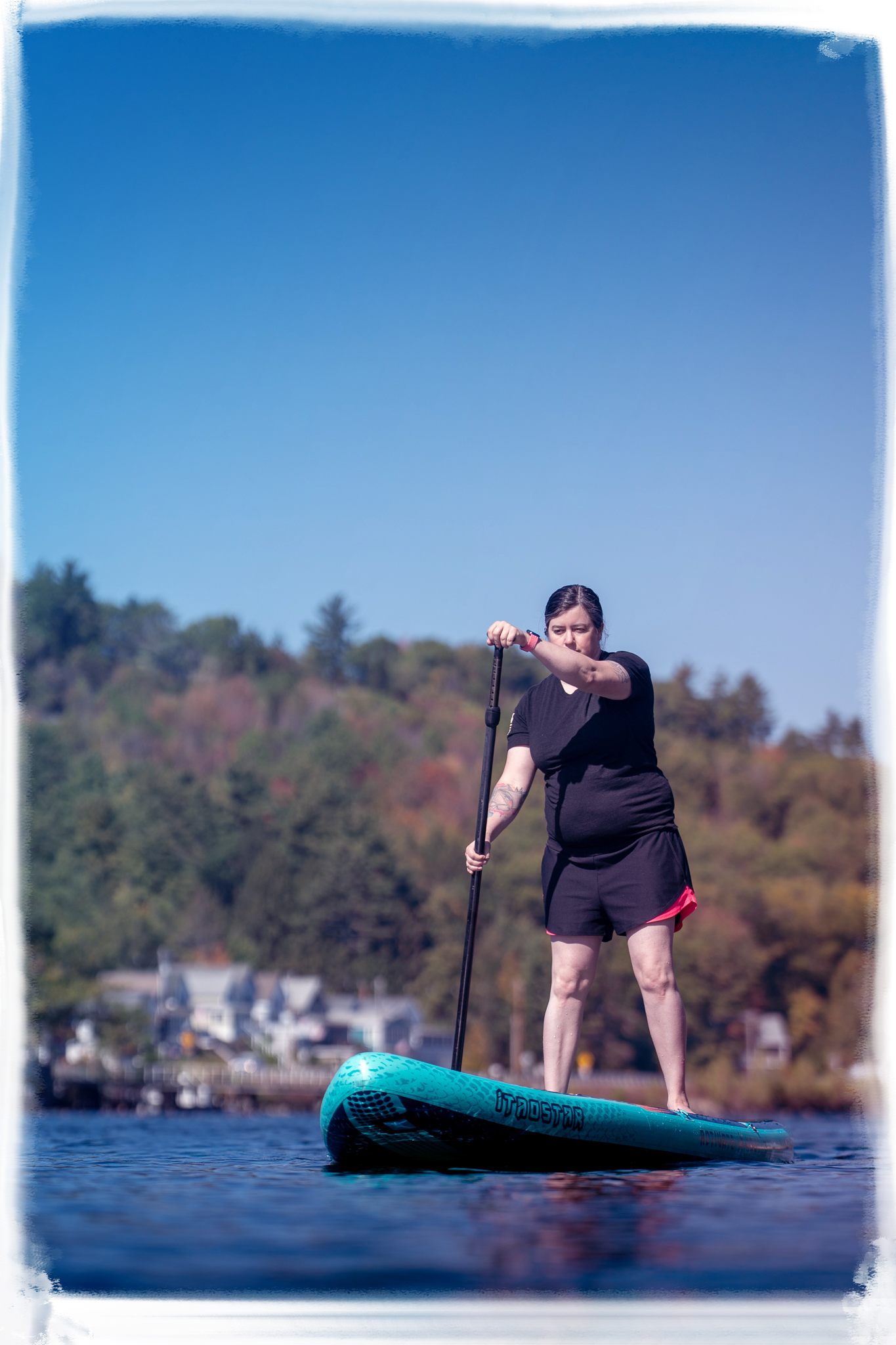 A person paddleboarding on calm blue waters near a wooded shoreline on a sunny day.