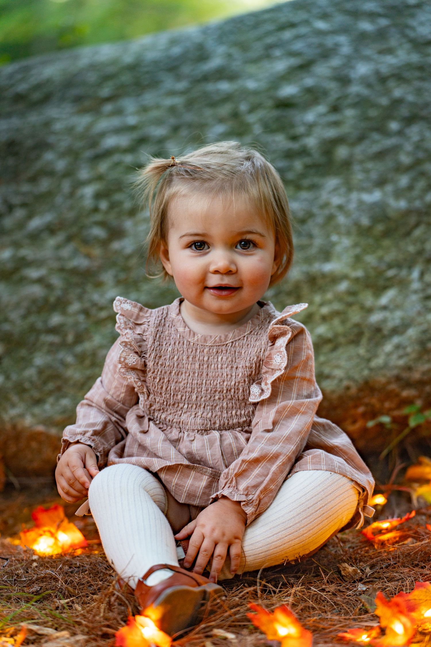 Baby in pink smocked dress sits on ground with autumn leaves and glowing string lights in background.