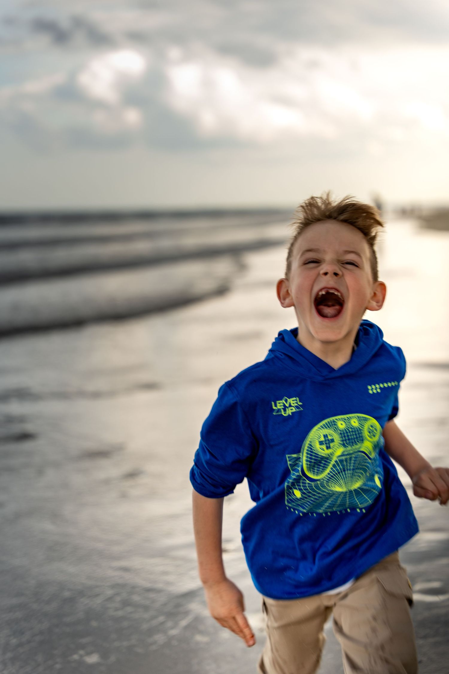 Excited young boy in blue gaming hoodie running on beach at sunset with mouth open wide.