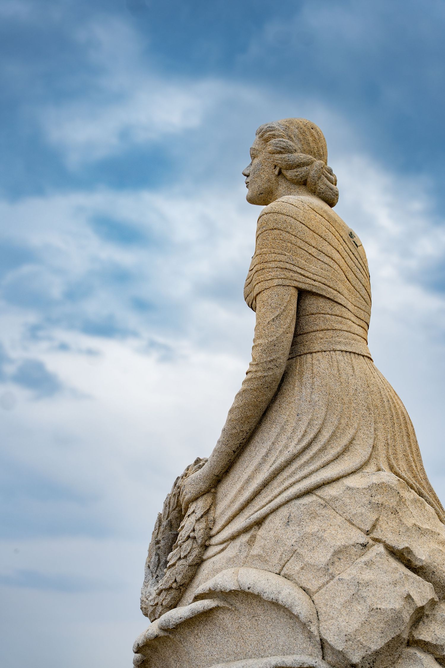 Stone statue of a woman in flowing dress and shawl, viewed from below against a cloudy blue sky.