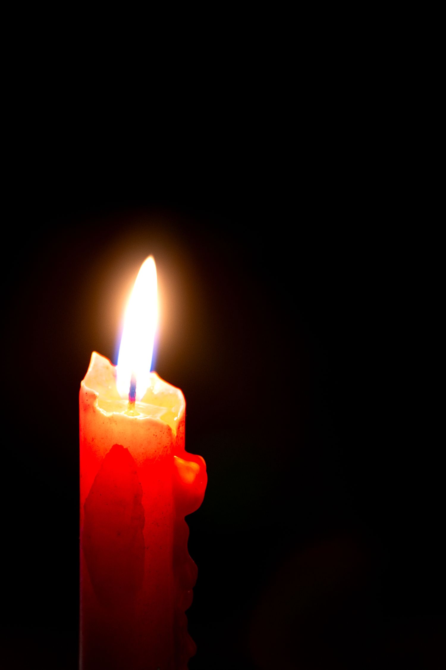 A glowing red candle with dripping wax burns brightly against a dark black background.
