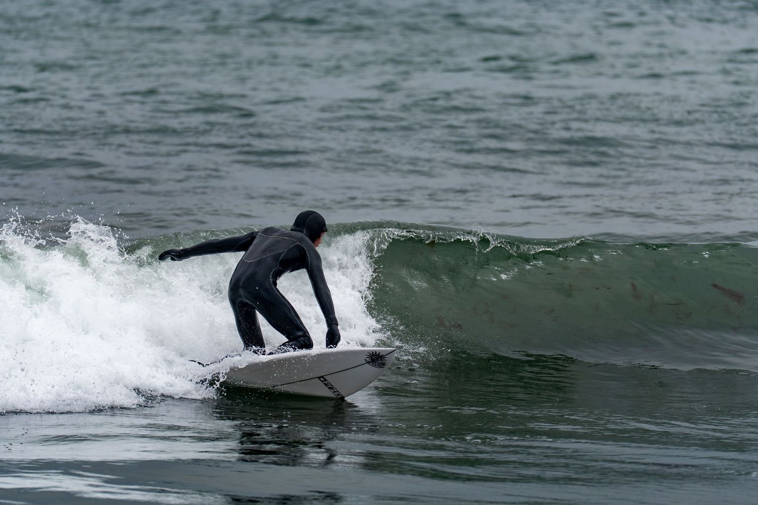 A surfer in a black wetsuit rides a wave on a white surfboard in cold ocean waters.