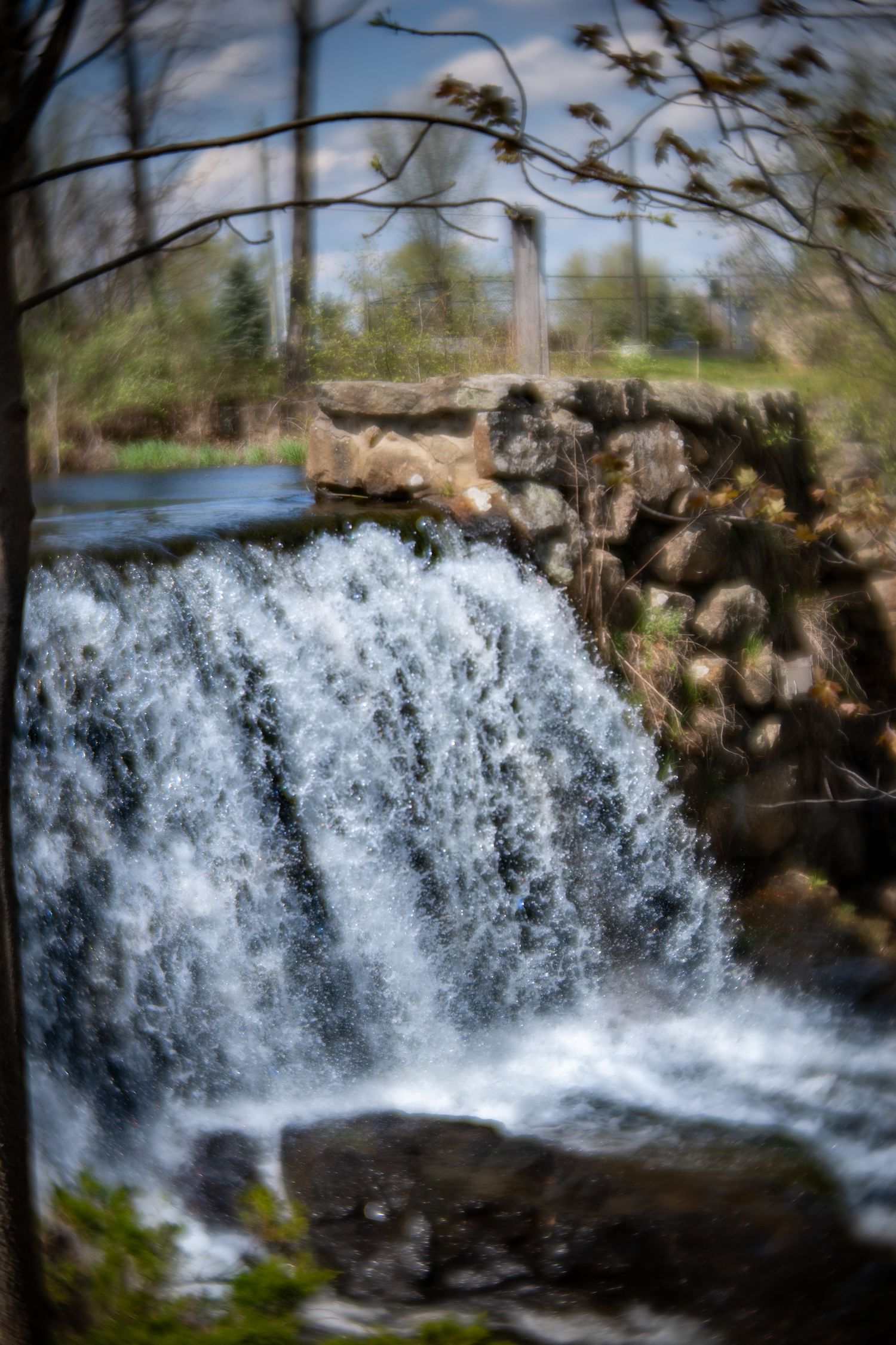 A rushing waterfall cascades over rocks beside a stone wall, framed by bare tree branches against a blue sky.