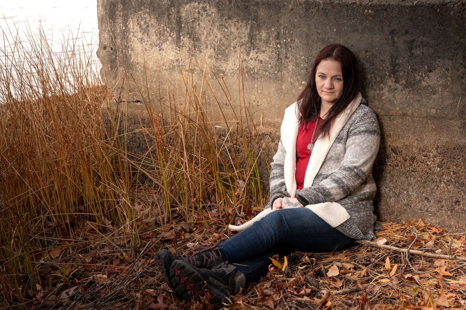Person in gray coat sits against rustic wall with autumn foliage in outdoor portrait session.
