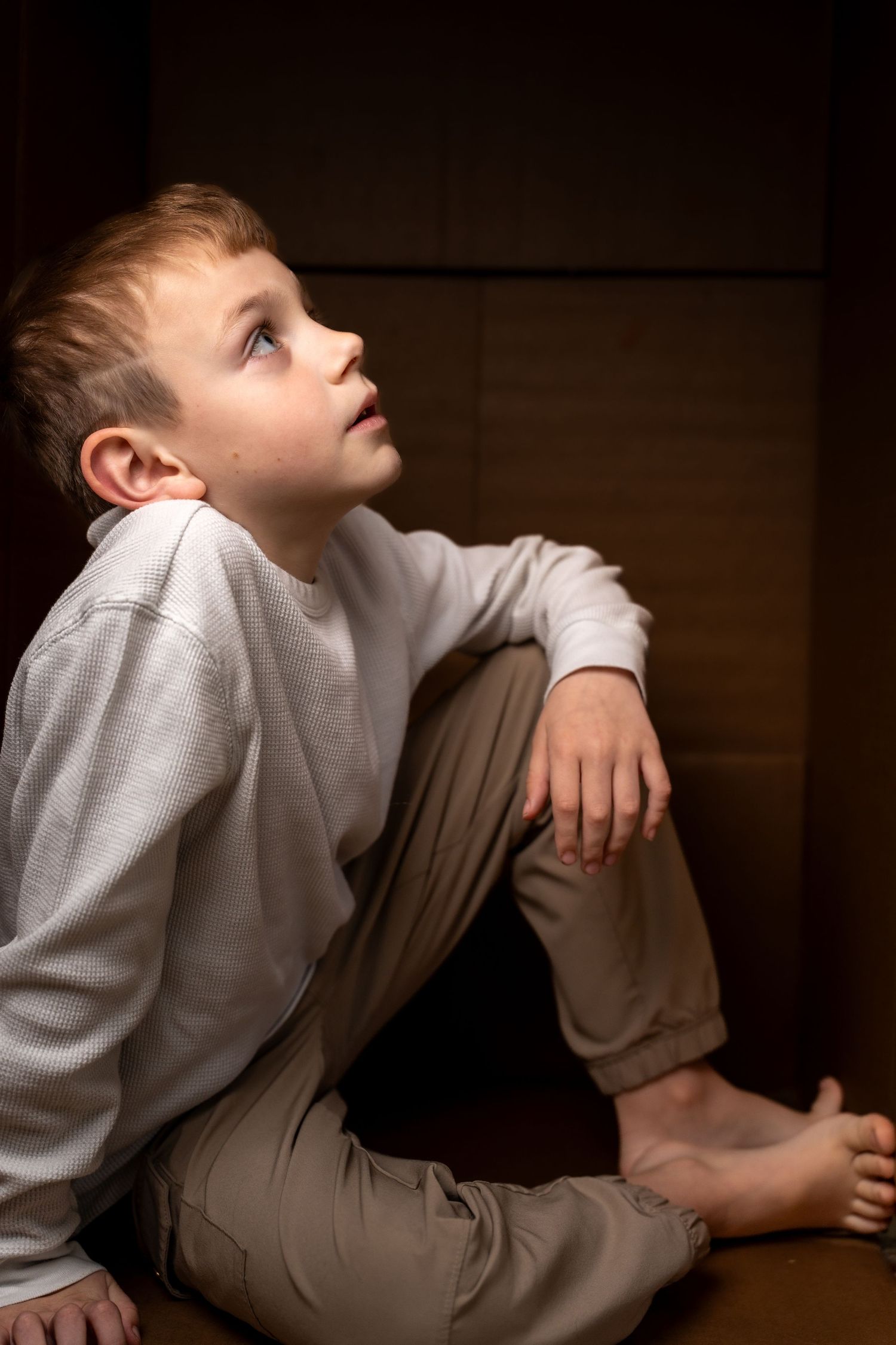 A barefoot boy in beige pants and white sweatshirt sits on floor in corner, gazing upward thoughtfully.