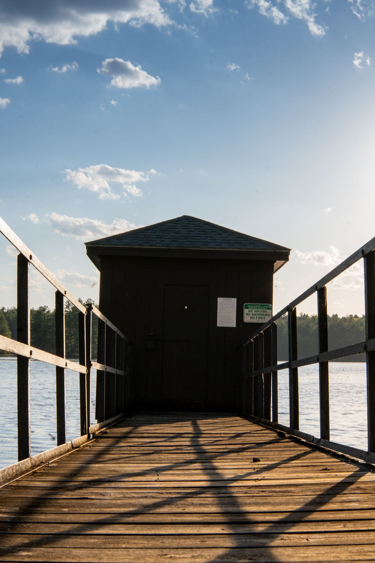 Wooden dock leading to a small dark boathouse over a calm lake under a partly cloudy blue sky.