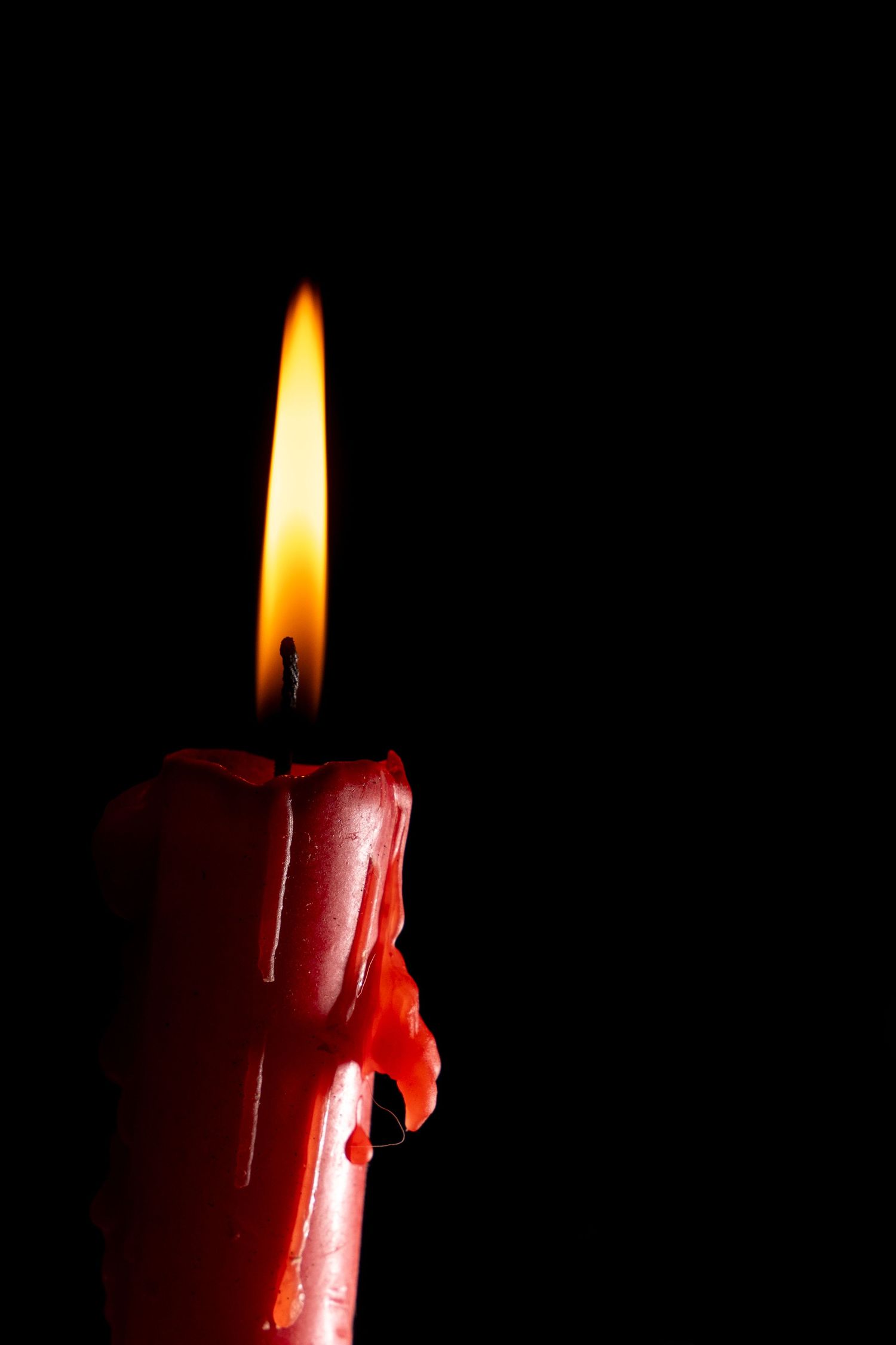 A red pillar candle with dripping wax burning with a bright flame against a dark black background.
