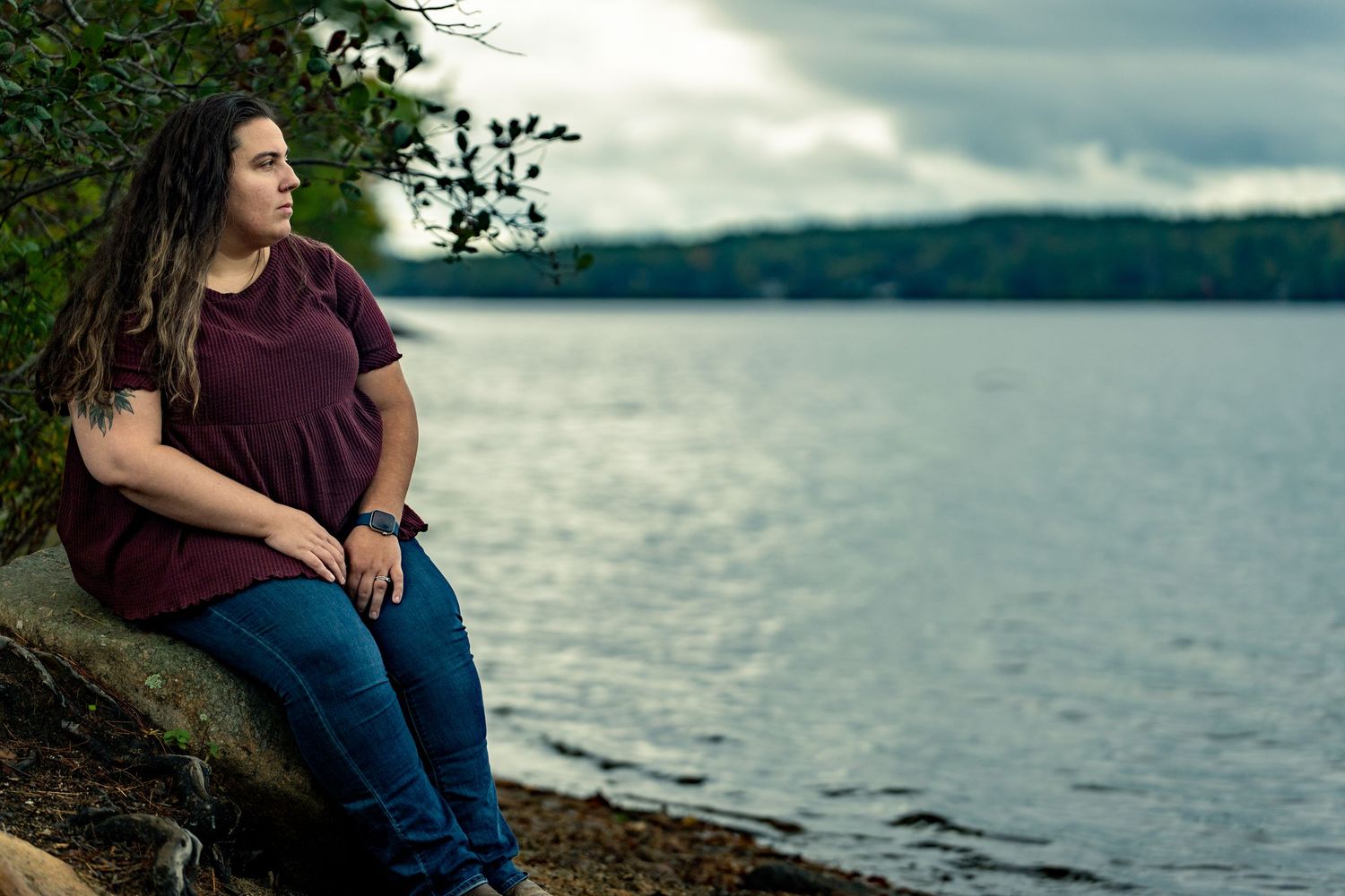 Person sits on rocky shoreline overlooking serene lake with mountains and cloudy sky in background.