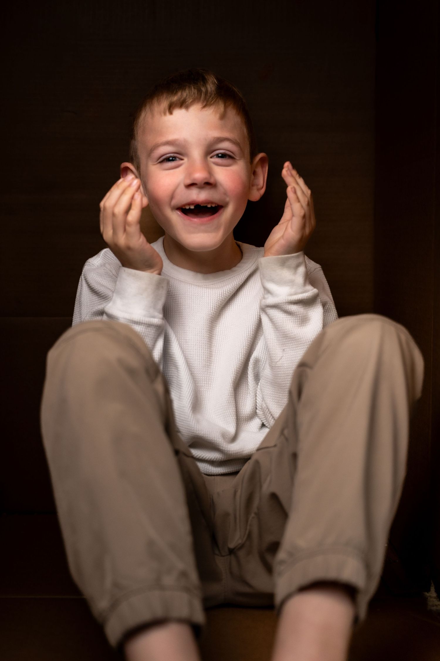 A happy young boy sitting on the floor laughing with hands raised beside his face, wearing a white sweatshirt and tan pants.