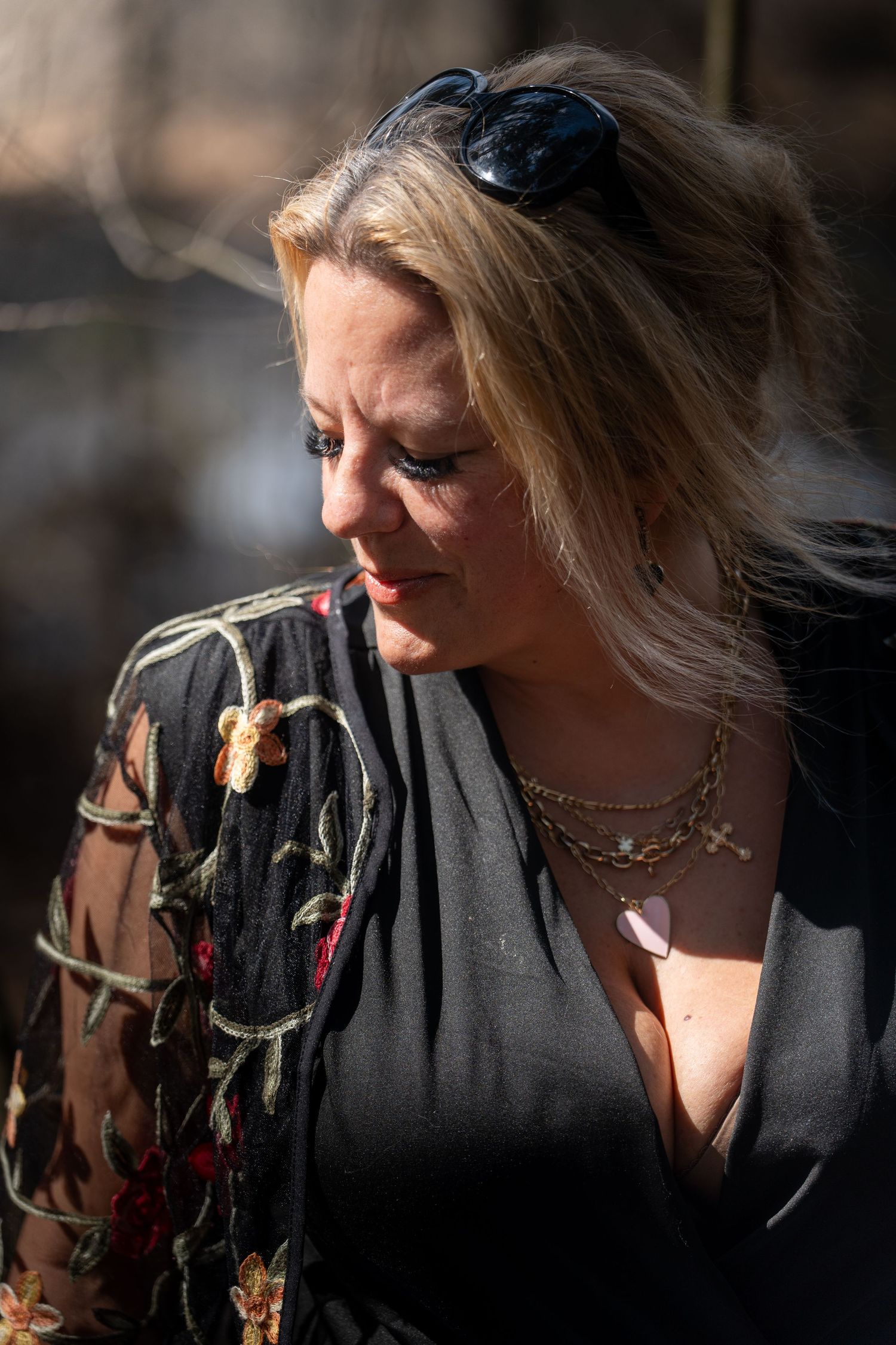 Blonde woman with sunglasses on head wearing floral embroidered black top and layered necklaces outdoors in sunlight.