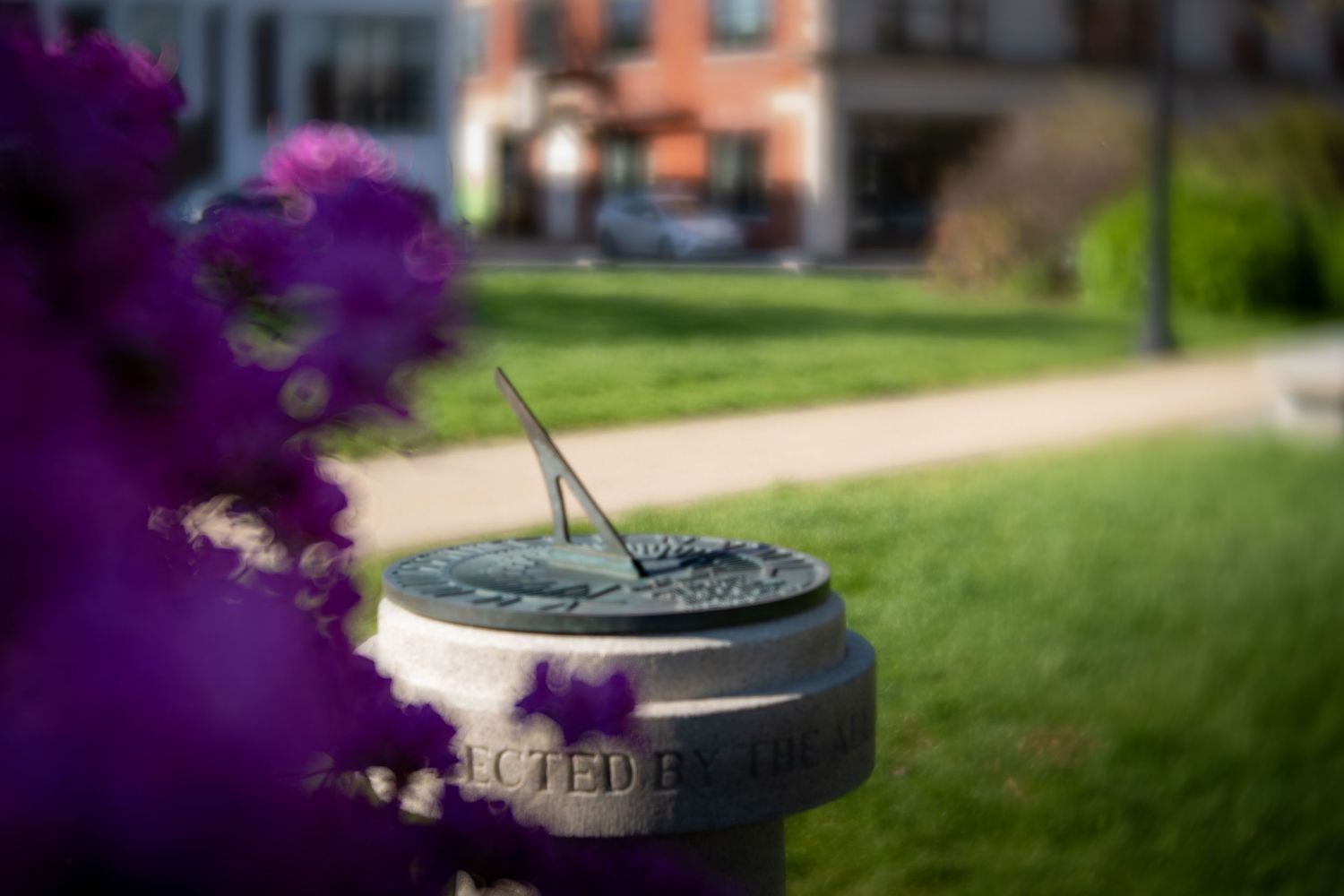 A sundial on a pedestal surrounded by purple smoke on a green lawn with a brick building in the background.