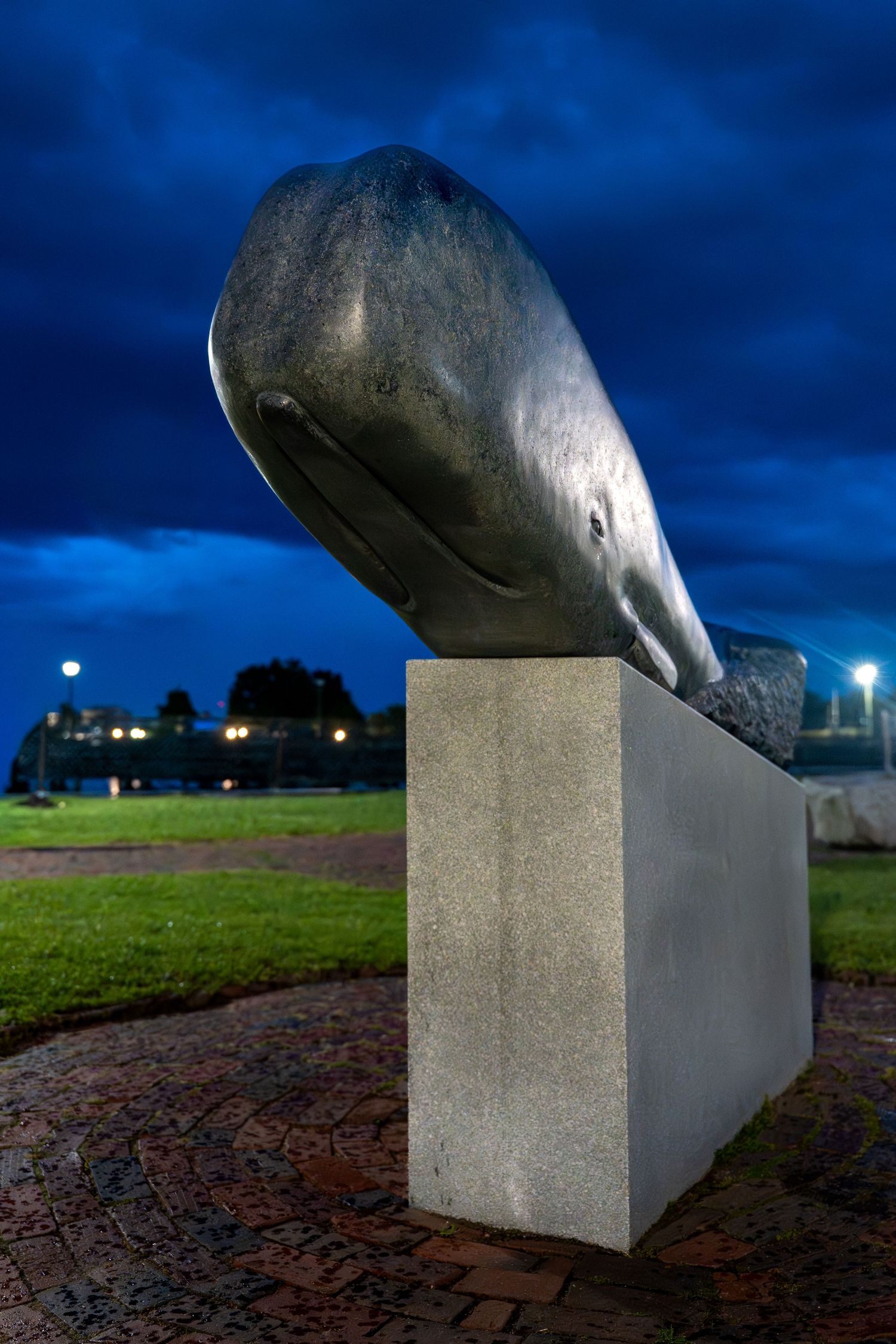 Bronze whale head sculpture on concrete pedestal at dusk, with dramatic blue cloudy sky and city lights in background.