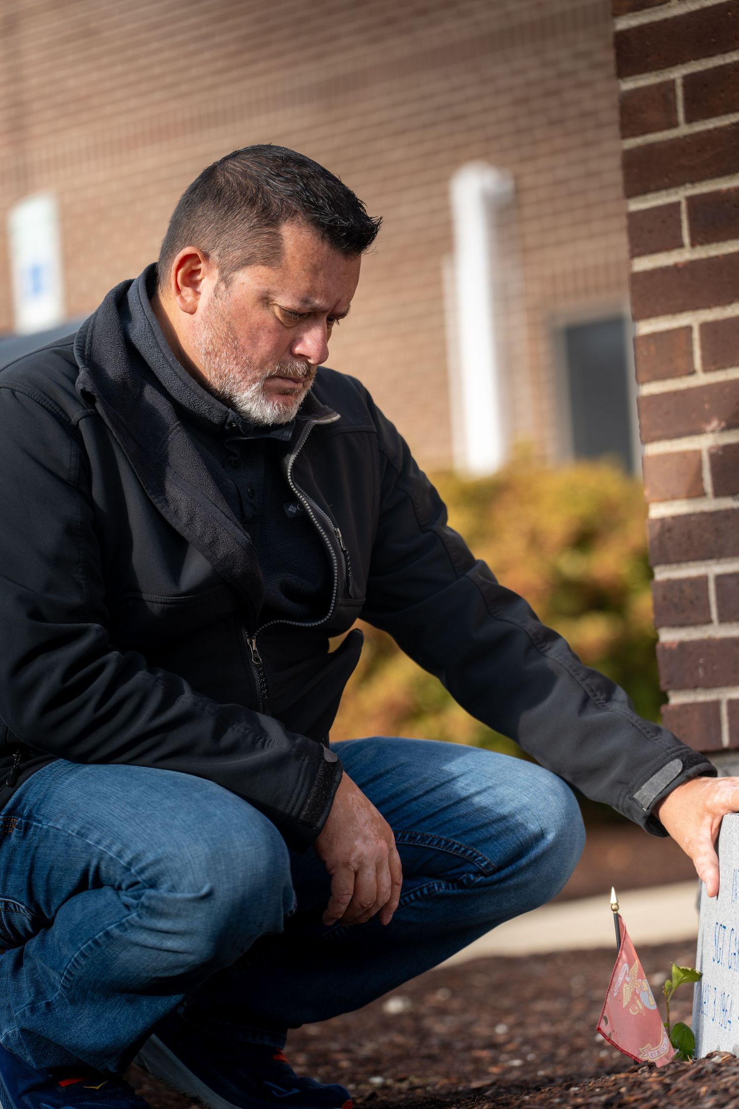 Person in black jacket and jeans kneels by brick wall while placing something on ground.