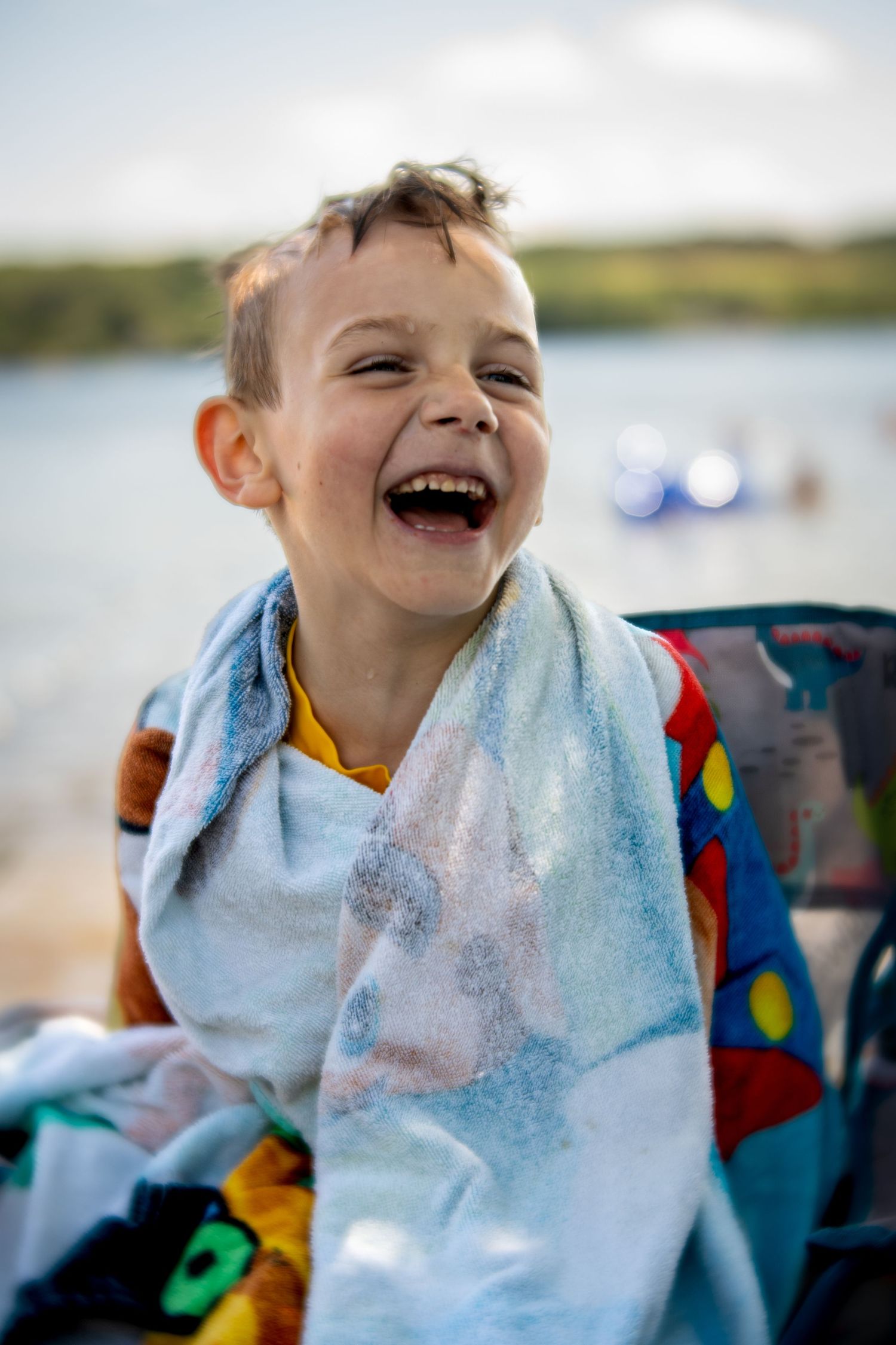 A laughing young boy wrapped in a light blue towel sits outdoors near a lake on a sunny day.