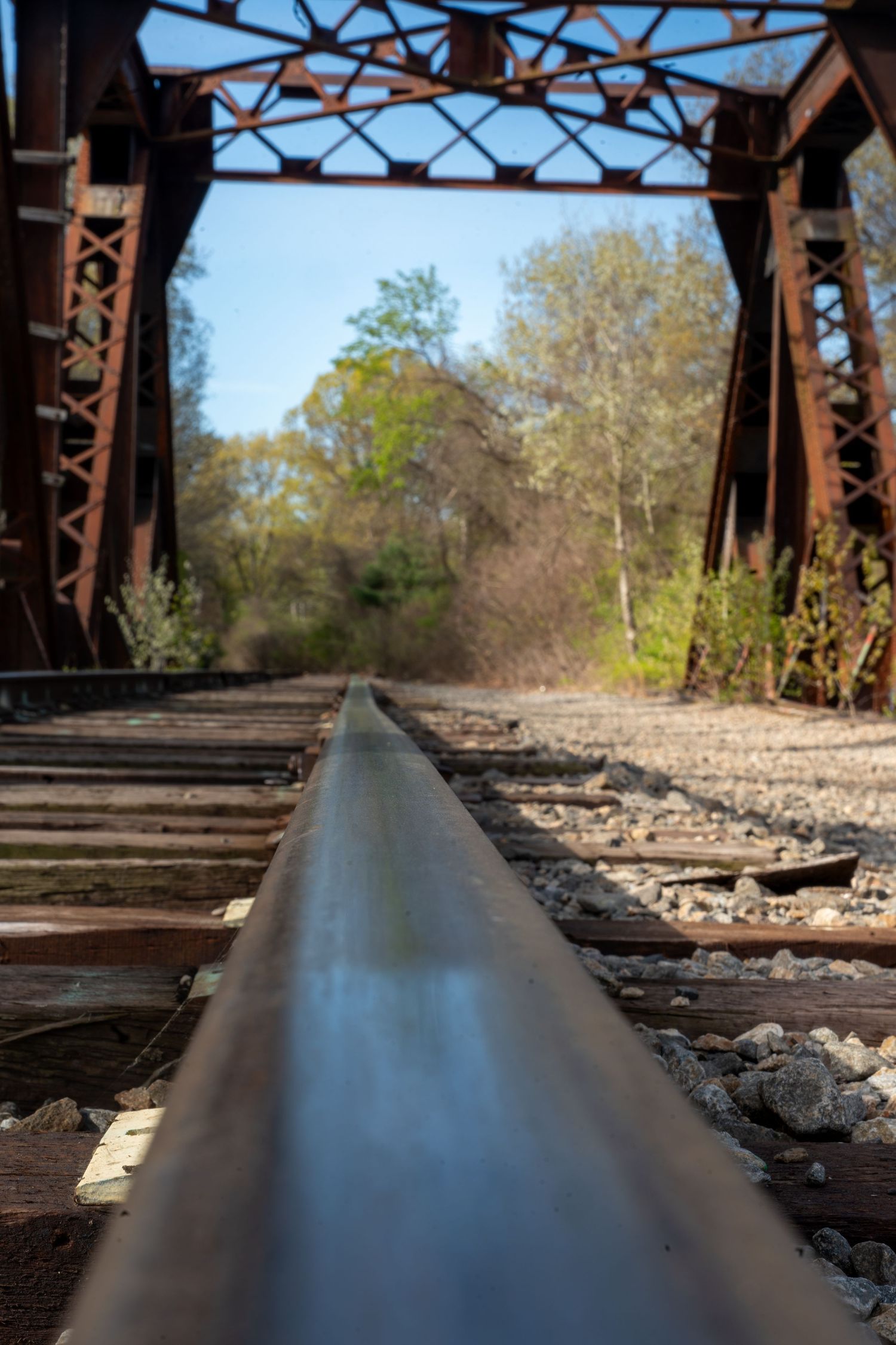 Low angle view of railroad tracks leading through a rusty iron trestle bridge surrounded by green trees.