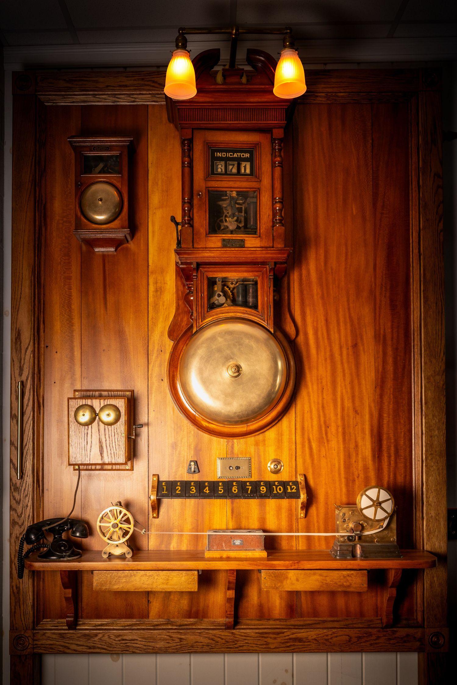 Vintage fire station alarm system with brass bell, indicator board, and antique telephone on wood wall.