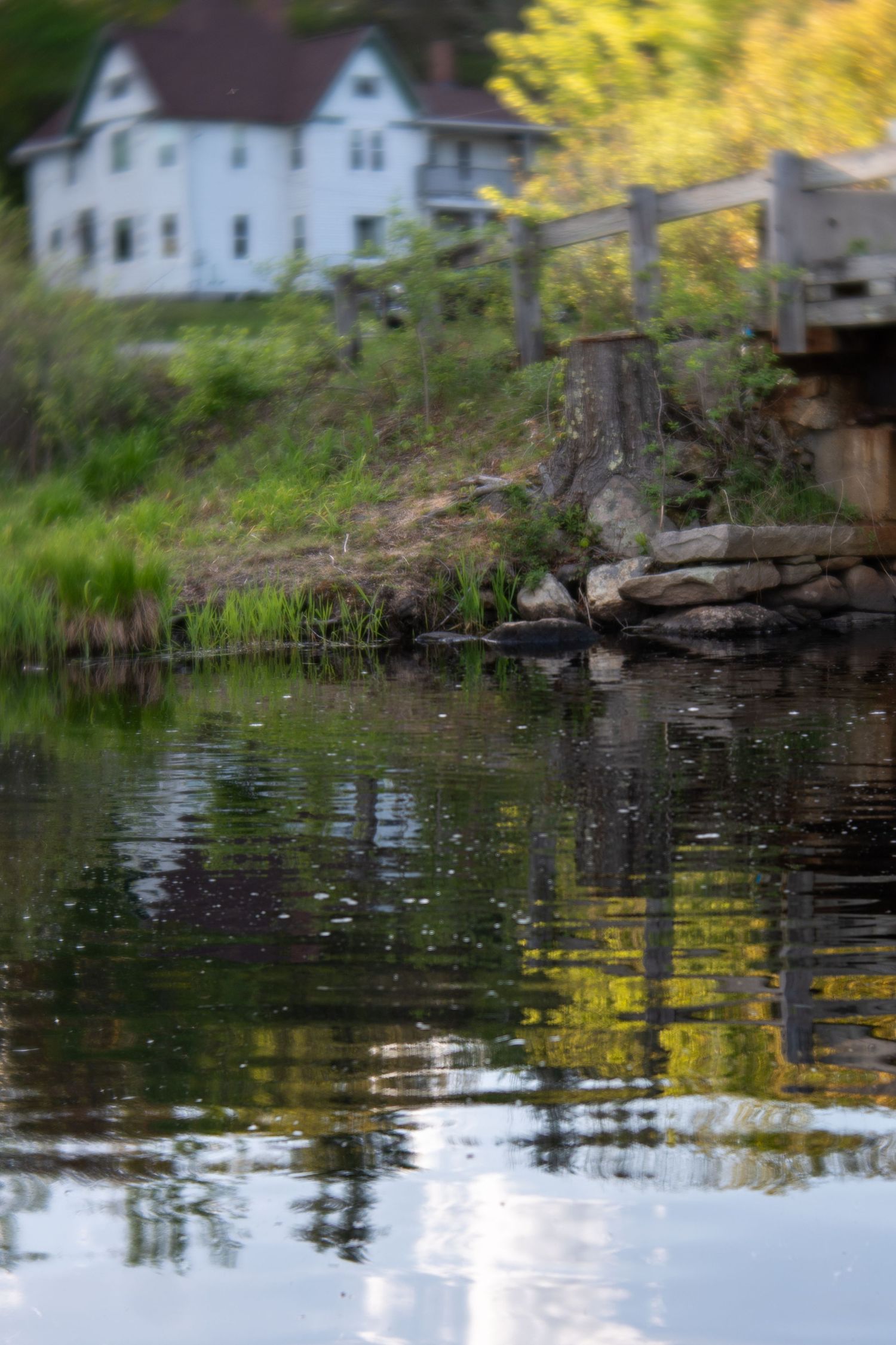 Calm river reflecting white house and green trees on rocky shoreline in a serene New England landscape.