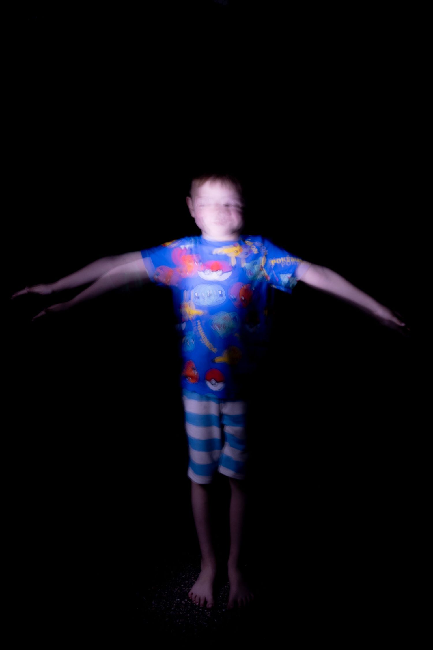 Blurry long exposure photo of a child in Pokemon pajamas standing with arms outstretched in a dark room.