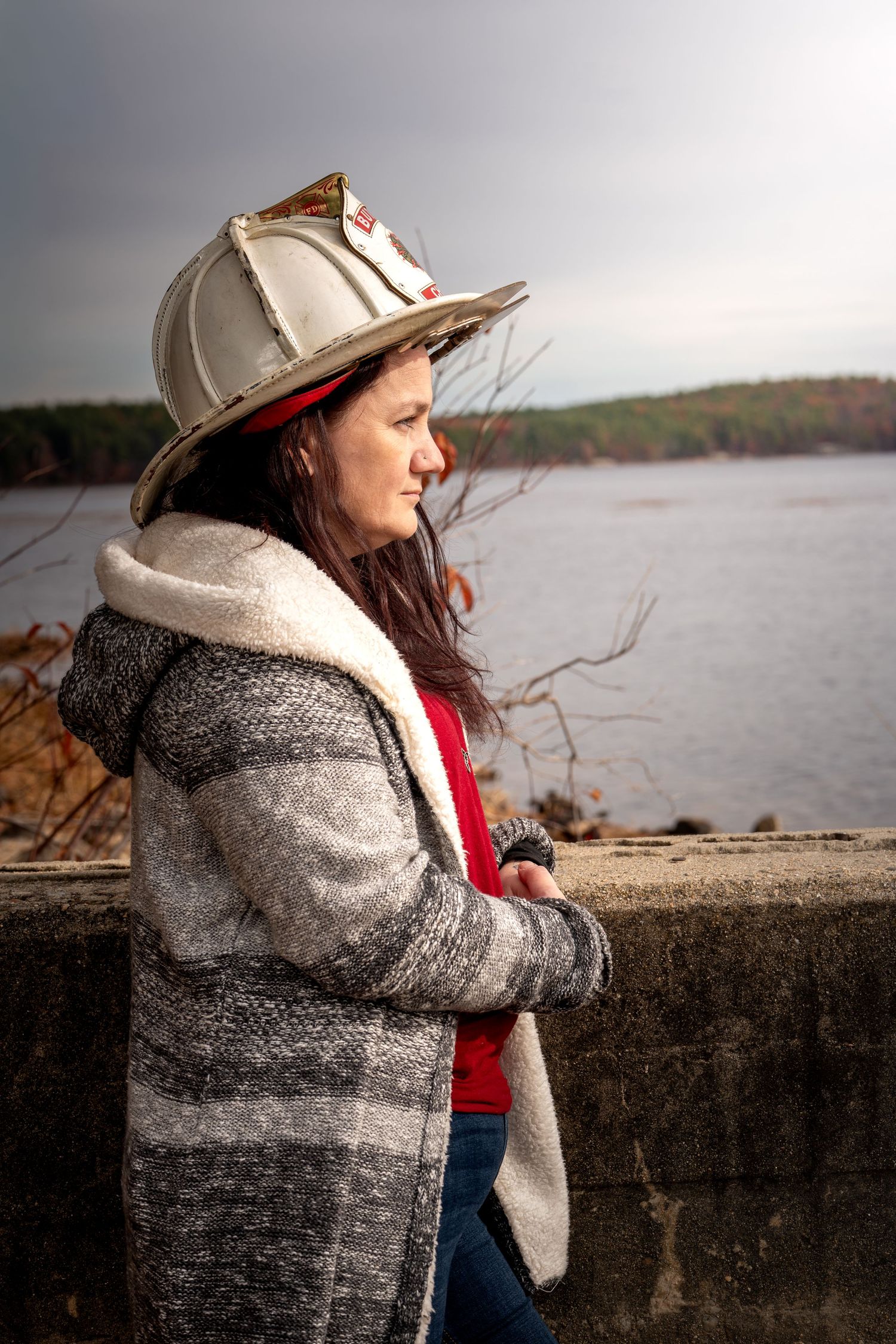 Person wearing a winter coat and baseball cap gazes thoughtfully at a scenic lake view on an overcast day.