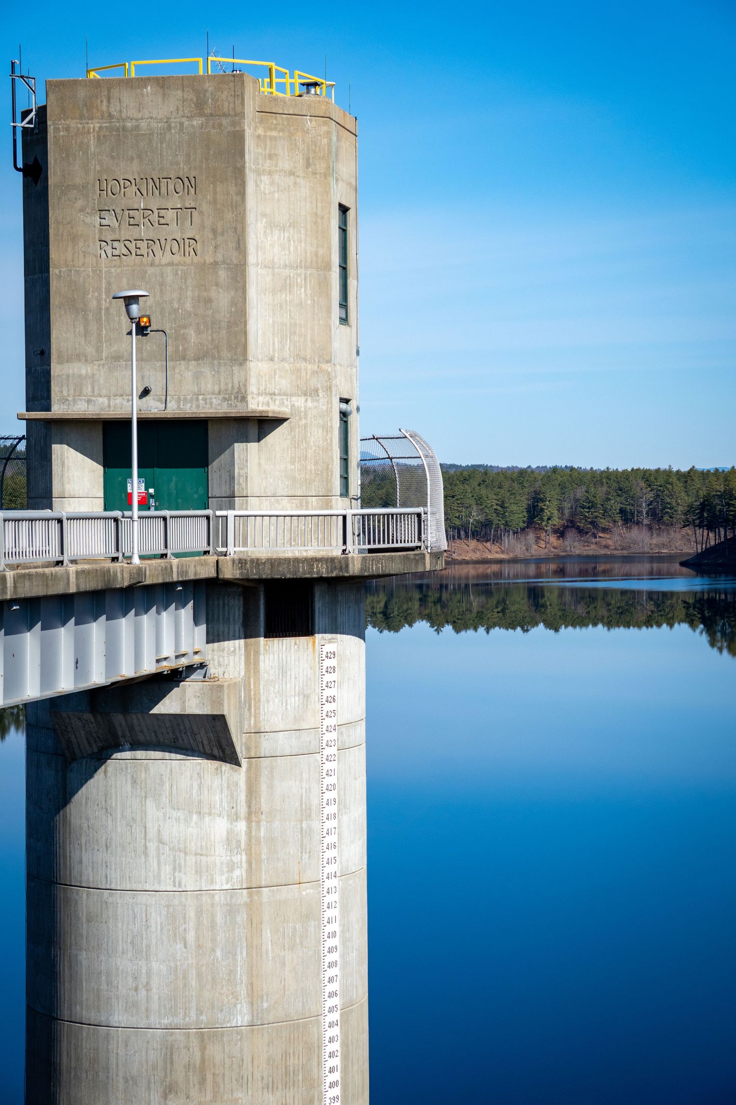 Concrete water reservoir towers with measurement markings stand against a calm blue lake and clear sky.