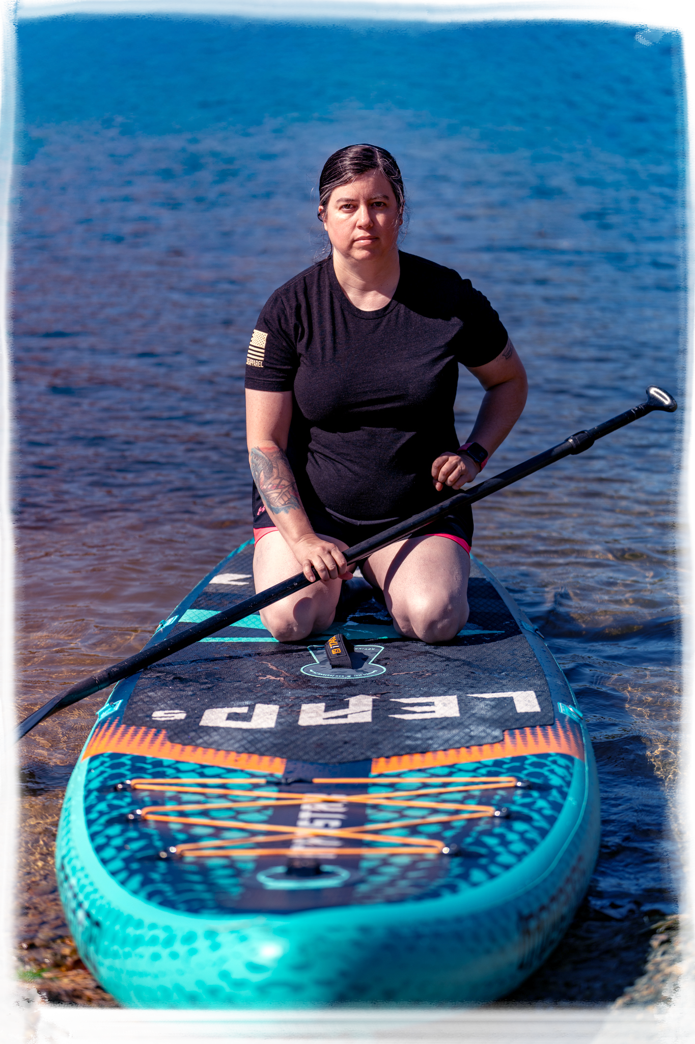 A person in black clothing sits on a turquoise paddleboard on calm blue waters.