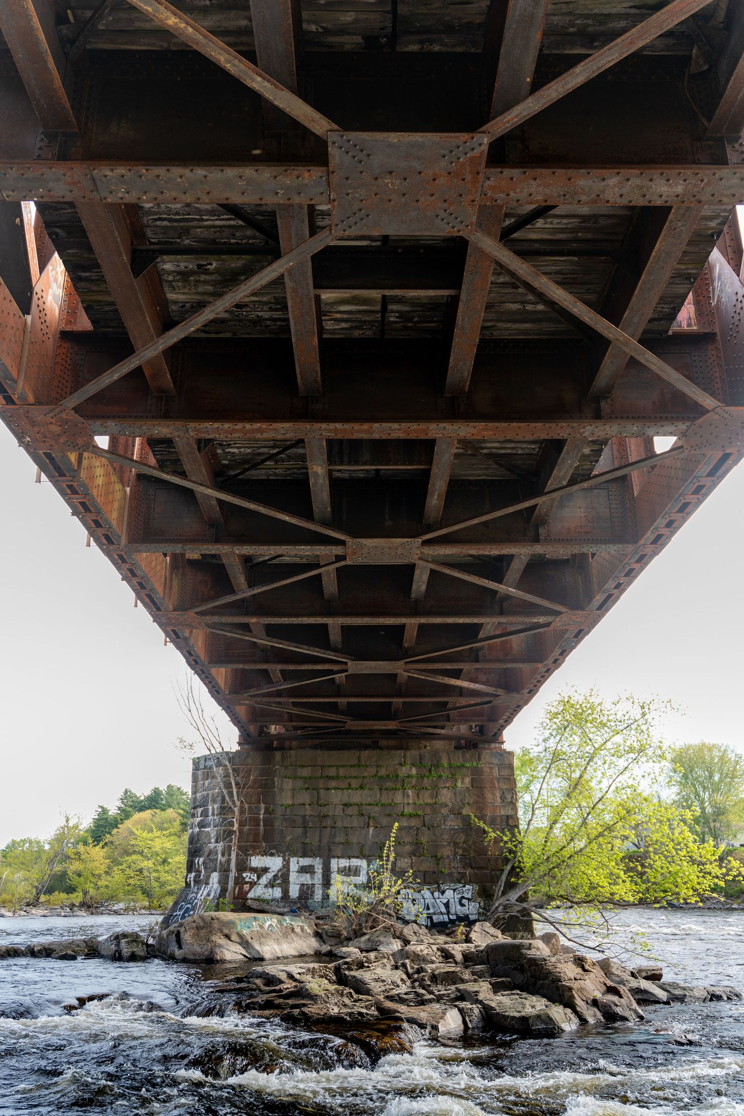 Rusty steel bridge underside with geometric truss structure over rocky river, graffiti reading ZAR on stone pillar, surrounded by trees.