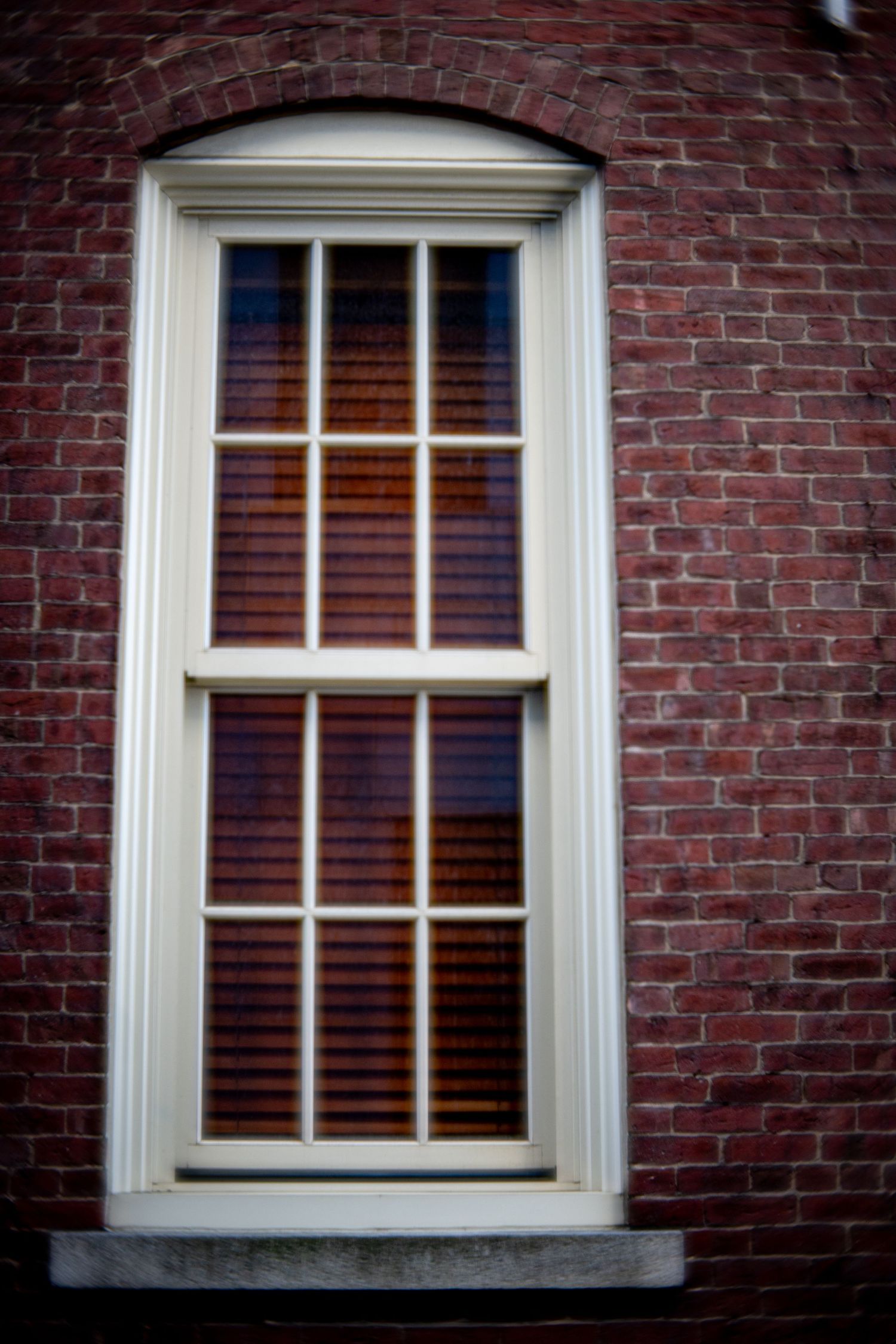 White-framed double-hung window with wooden blinds set in a classic red brick wall with an arched brick detail above.