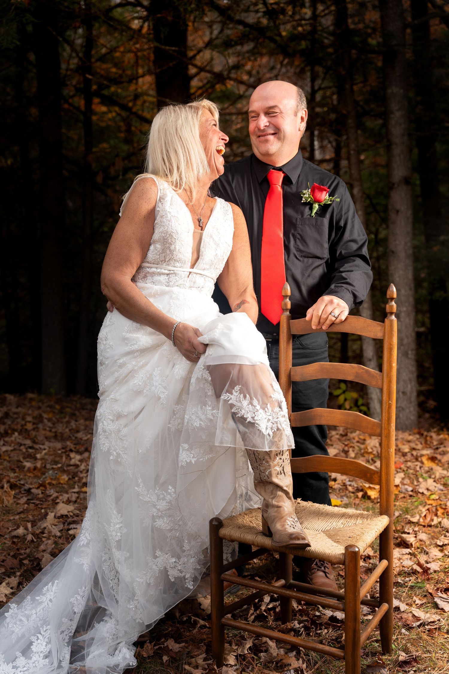 A couple shares joyful moments in autumn woods while sitting on rustic wooden chairs surrounded by fallen leaves.