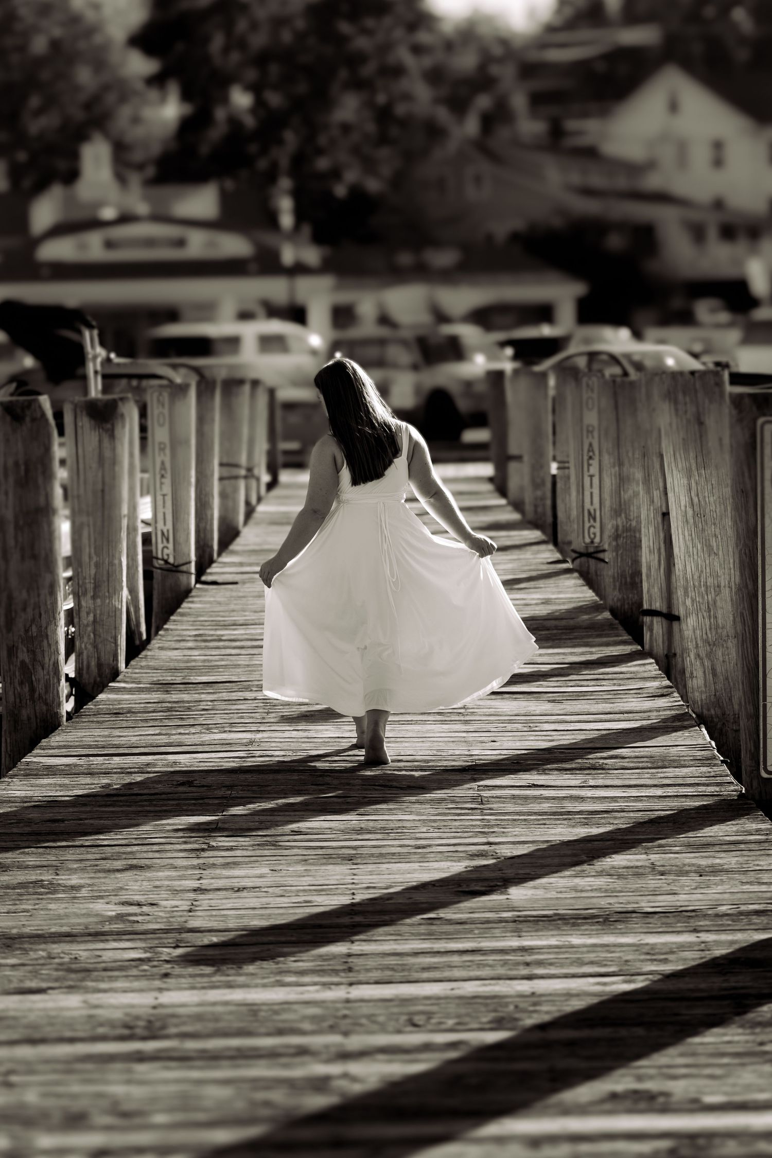 Black and white photo of a woman in a flowing white dress walking along a wooden marina dock.