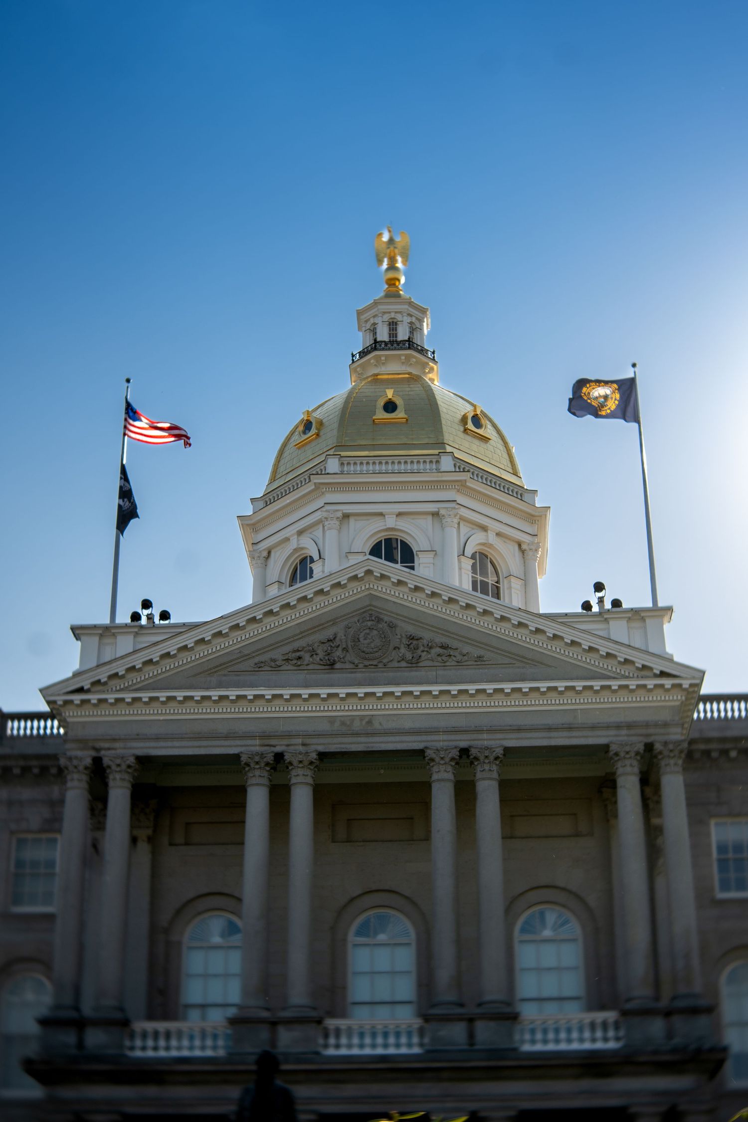 New Hampshire State Capitol building with gold dome, eagle statue, and three flags against a clear blue sky.
