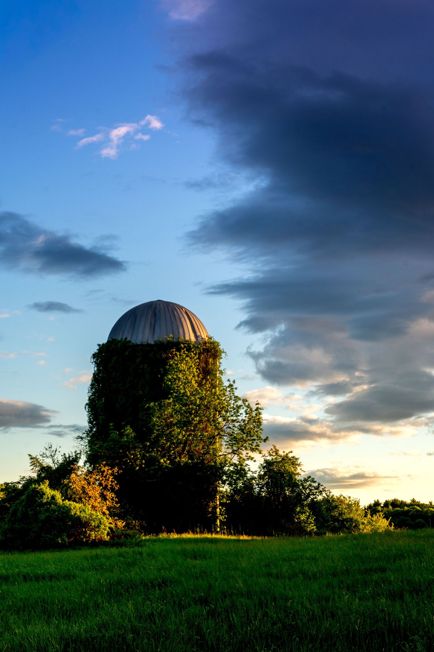 Abandoned silo covered in ivy stands in a green field under a dramatic stormy sunset sky.