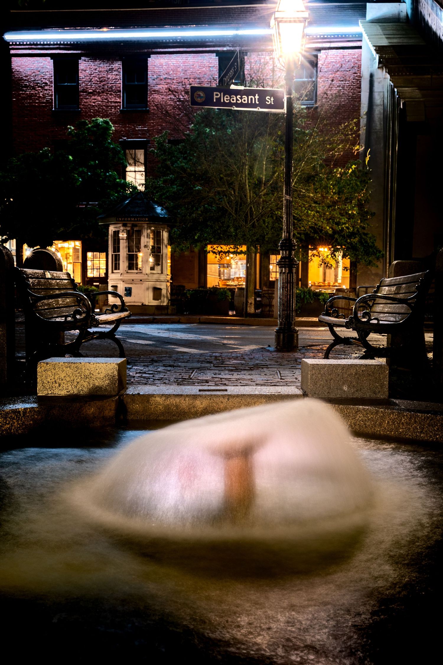 Snow-dusted fountain on Pleasant Street plaza at night with benches and glowing storefronts.