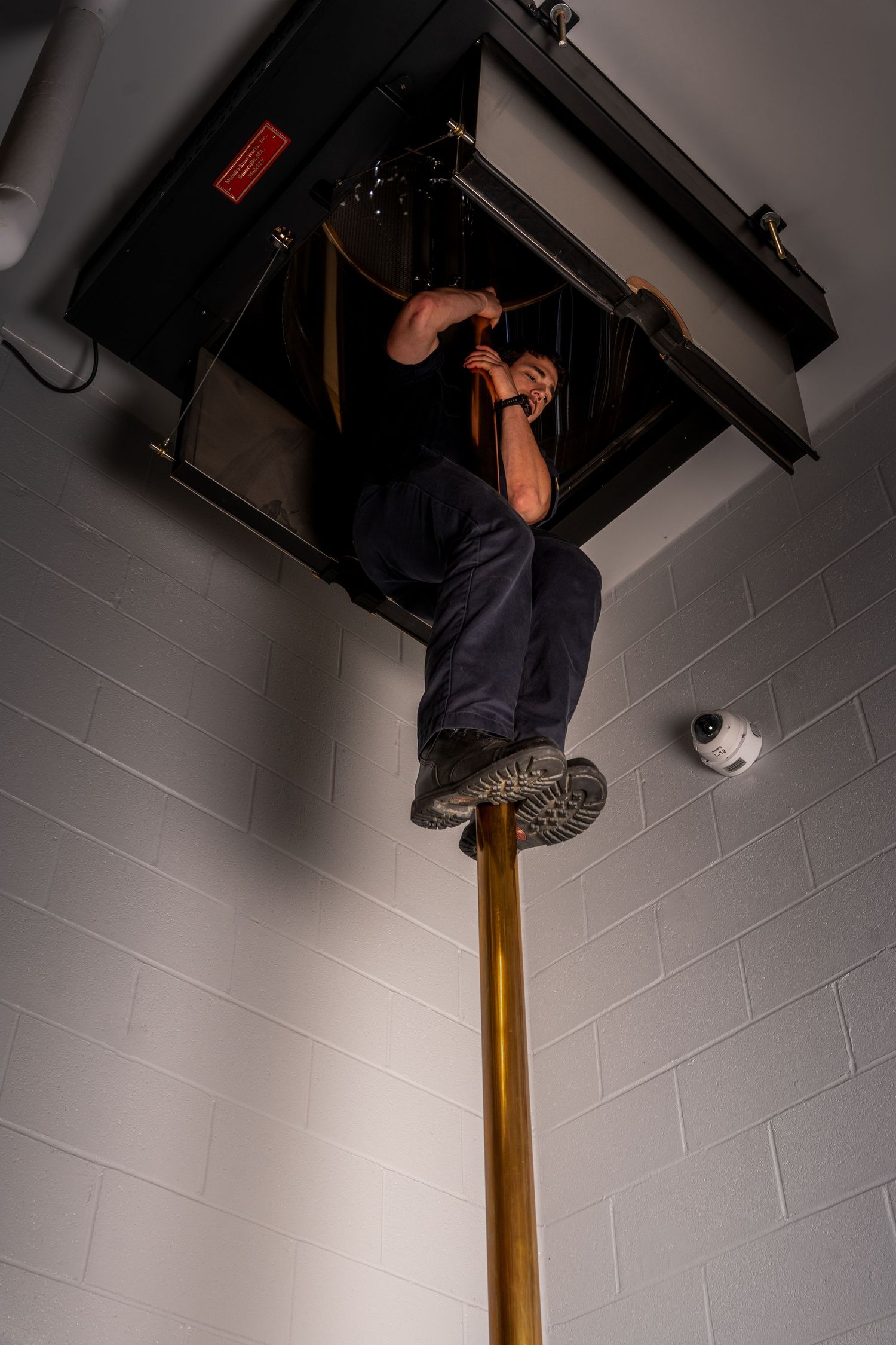 Technician climbing a pole to perform maintenance work on ceiling-mounted HVAC equipment in a white tiled room.