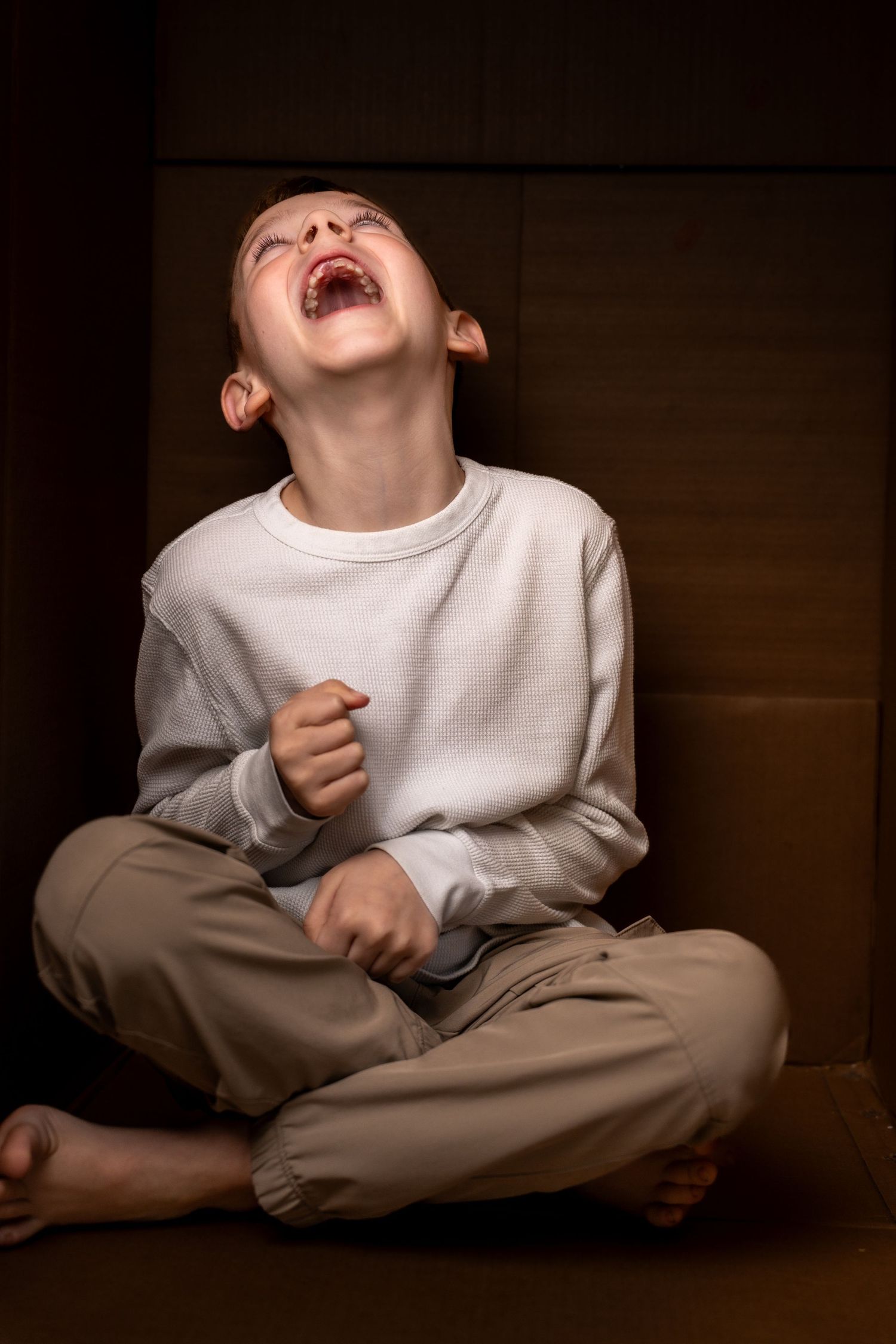 Young boy sitting cross-legged on floor, laughing with head tilted back, wearing white sweatshirt and tan pants.