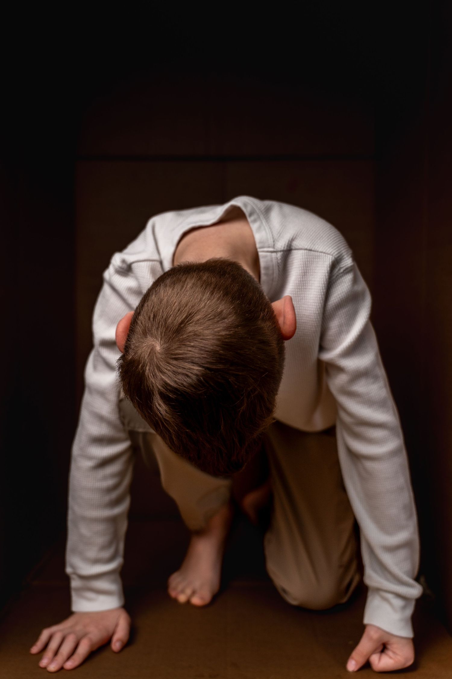 A child in a white outfit kneeling on all fours with head bowed down in a dark room.