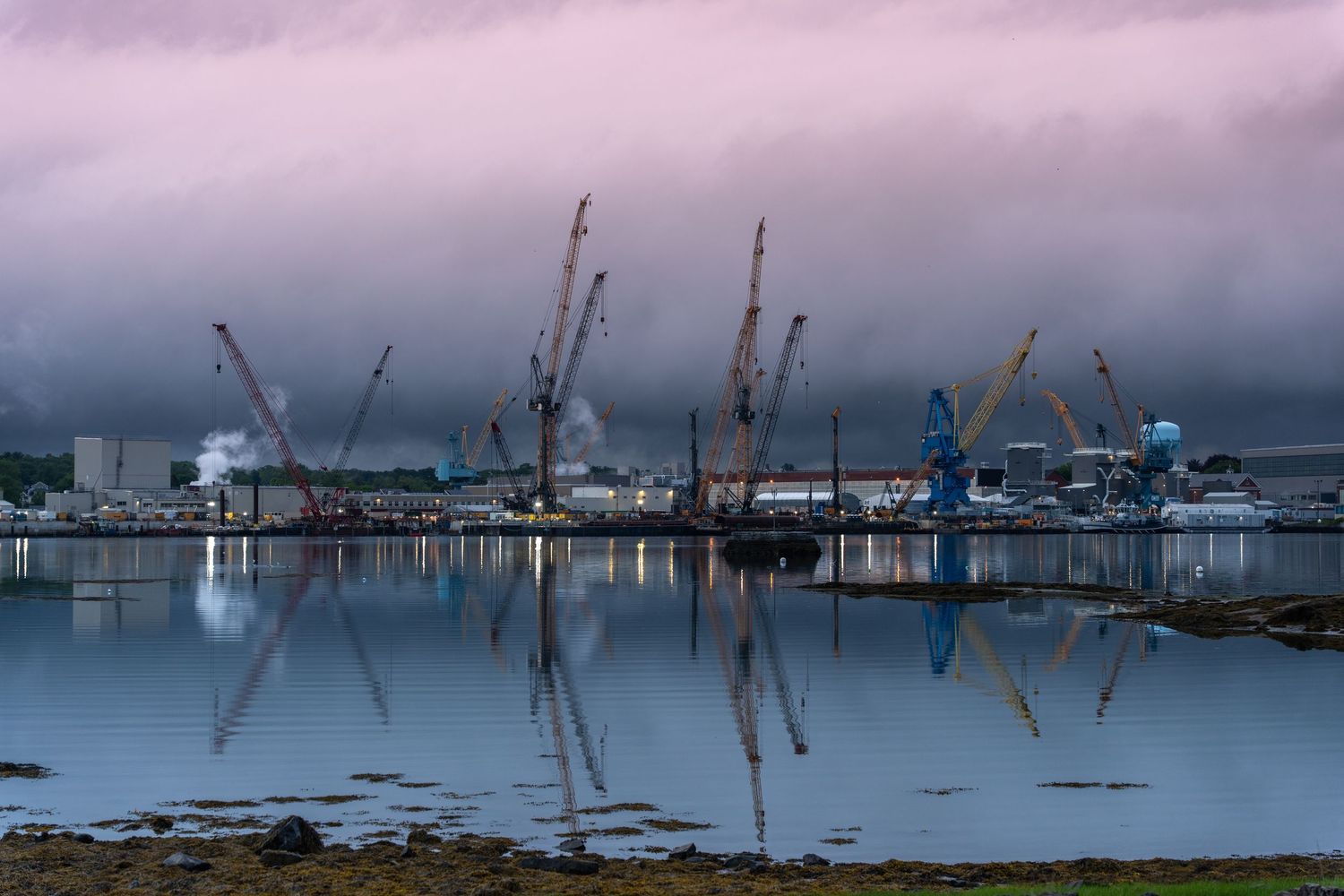 Industrial port cranes reflect on calm harbor waters at dusk under dramatic pink and purple storm clouds.