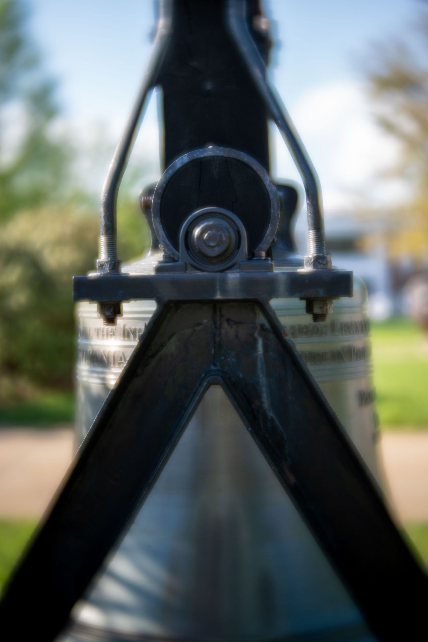 Close-up of a metal bike frame junction with bolts and V-shaped support, blurred green park background.